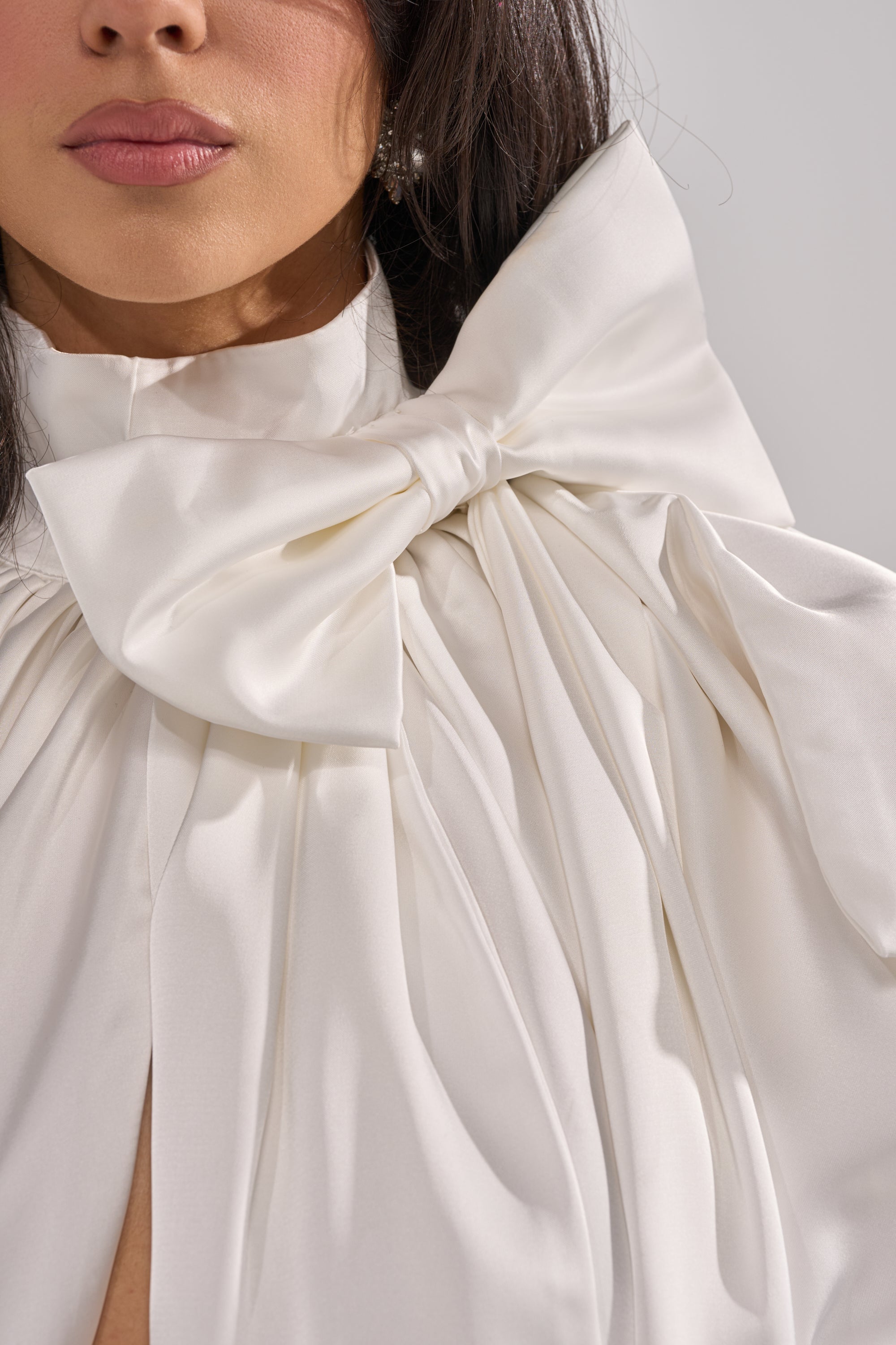 A woman wears the WRAPPED IN LOVE BLOUSE—an elegant white organza top with a sculptural shoulder bow, soft pleats, and flowing fabric. Only her lower face and upper torso are shown. 6