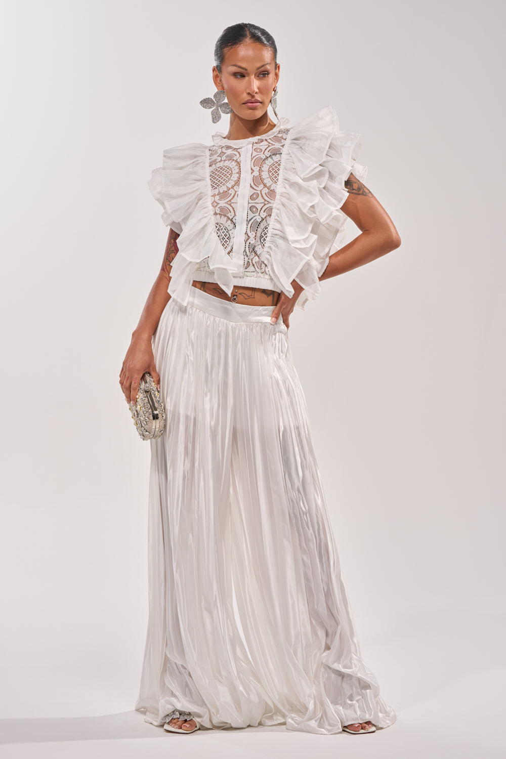 A woman models the CALLED OUT SATIN PLEATED PALAZZO PANT IN WHITE, featuring wide-leg satin pleats. She pairs them with a ruffled lace top, a small clutch, and bold earrings while posing confidently against a plain background. 1