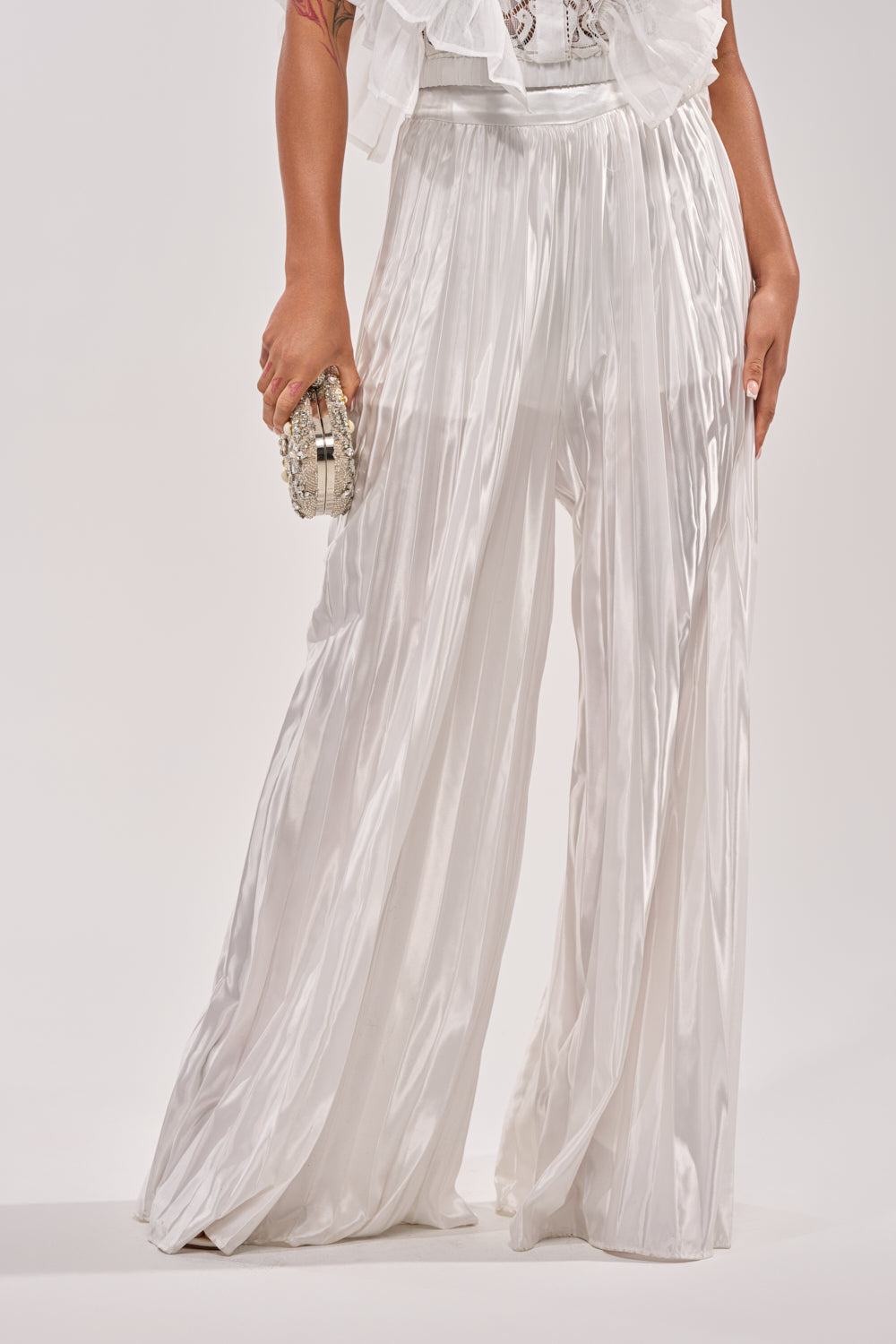 Wearing the CALLED OUT SATIN PLEATED PALAZZO PANT IN WHITE, a person stands against a light background holding a small textured clutch, with only the lower half of their body visible. 2