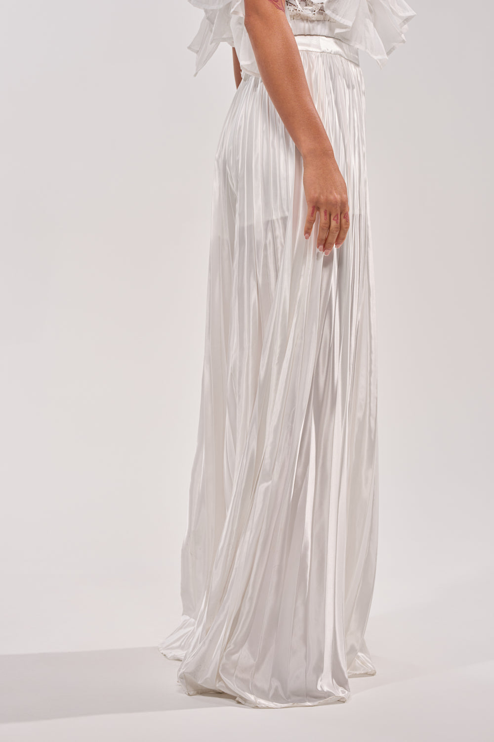 Wearing the CALLED OUT SATIN PLEATED PALAZZO PANT IN WHITE, a person stands against a plain light background, with only their lower half visible and an arm resting by their side. The wide-leg pants feature a sheer, shiny satin fabric. 3