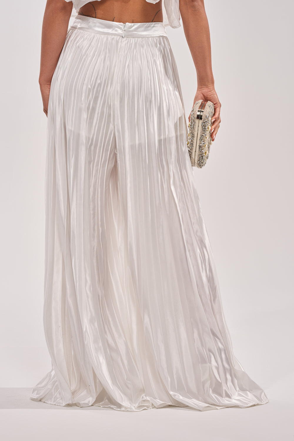 A person is seen from the back wearing the CALLED OUT SATIN PLEATED PALAZZO PANT IN WHITE, holding a small embellished clutch against a plain light background. 4