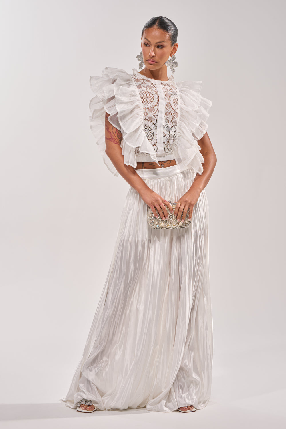 A woman models the CALLED OUT SATIN PLEATED PALAZZO PANT IN WHITE with a ruffled lace top, silver embellished clutch, statement earrings, and silver heels—an ideal look for chic office style. 6