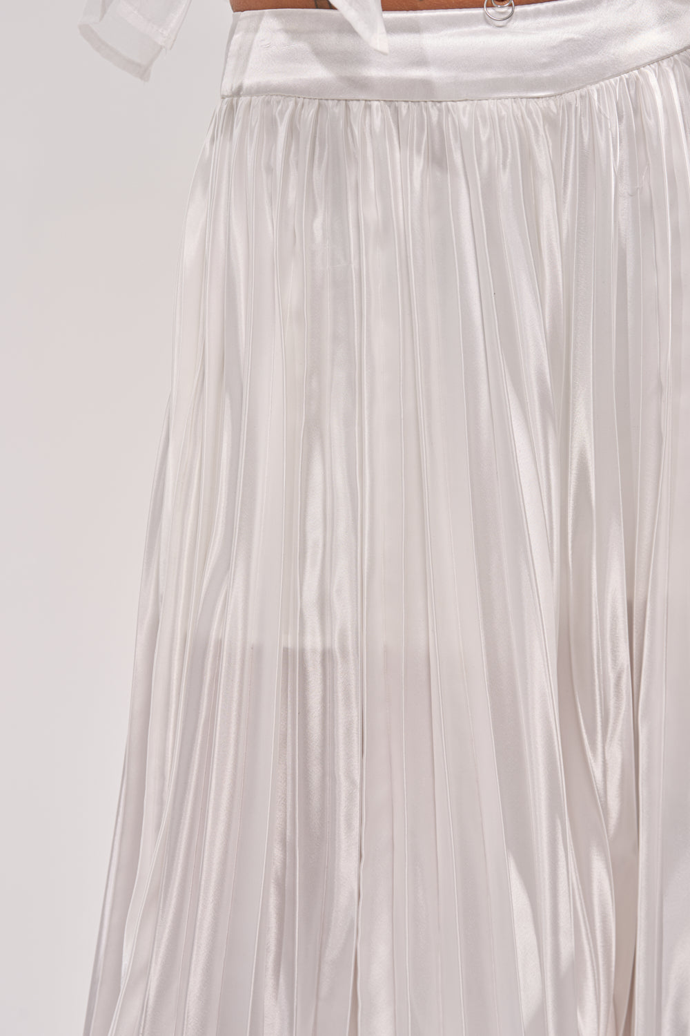 Close-up of CALLED OUT SATIN PLEATED PALAZZO PANT IN WHITE, featuring white shiny pleated satin, a silky waistband, and soft flowing texture. The high-rise wide-leg design offers elegant style for the office against a plain, light-colored background. 7