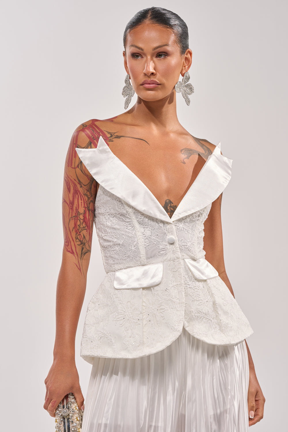 A woman with tattoos wears the CALLED OUT LACE CORSET, a white textured deep V-neck lace overlay top with lapels and peplum details, paired with a pleated skirt. She accessorizes with statement earrings and holds a sparkling clutch. 0
