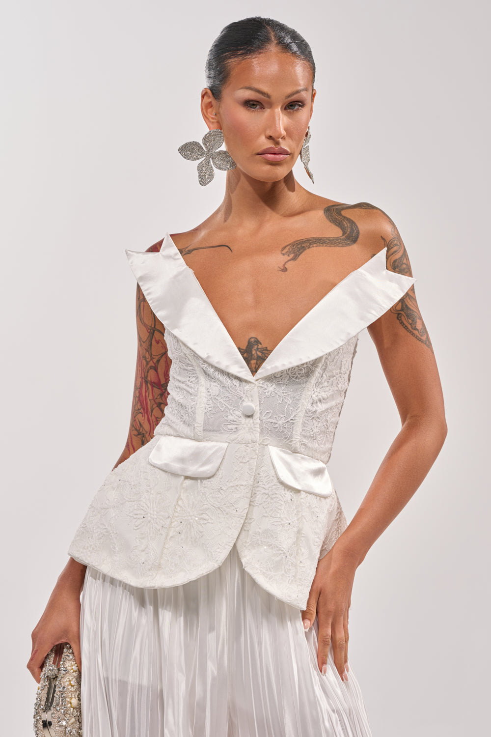 A woman wears the CALLED OUT LACE CORSET—a white, off-the-shoulder top with ruffles and lace details—paired with a white pleated skirt, large floral earrings, and visible arm and chest tattoos. 1