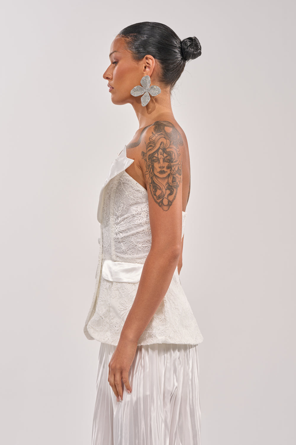 A woman in profile poses against a plain background, wearing the CALLED OUT LACE CORSET—a textured white strapless top—paired with large silver flower earrings, her hair in a sleek bun, and a detailed upper arm tattoo. 2