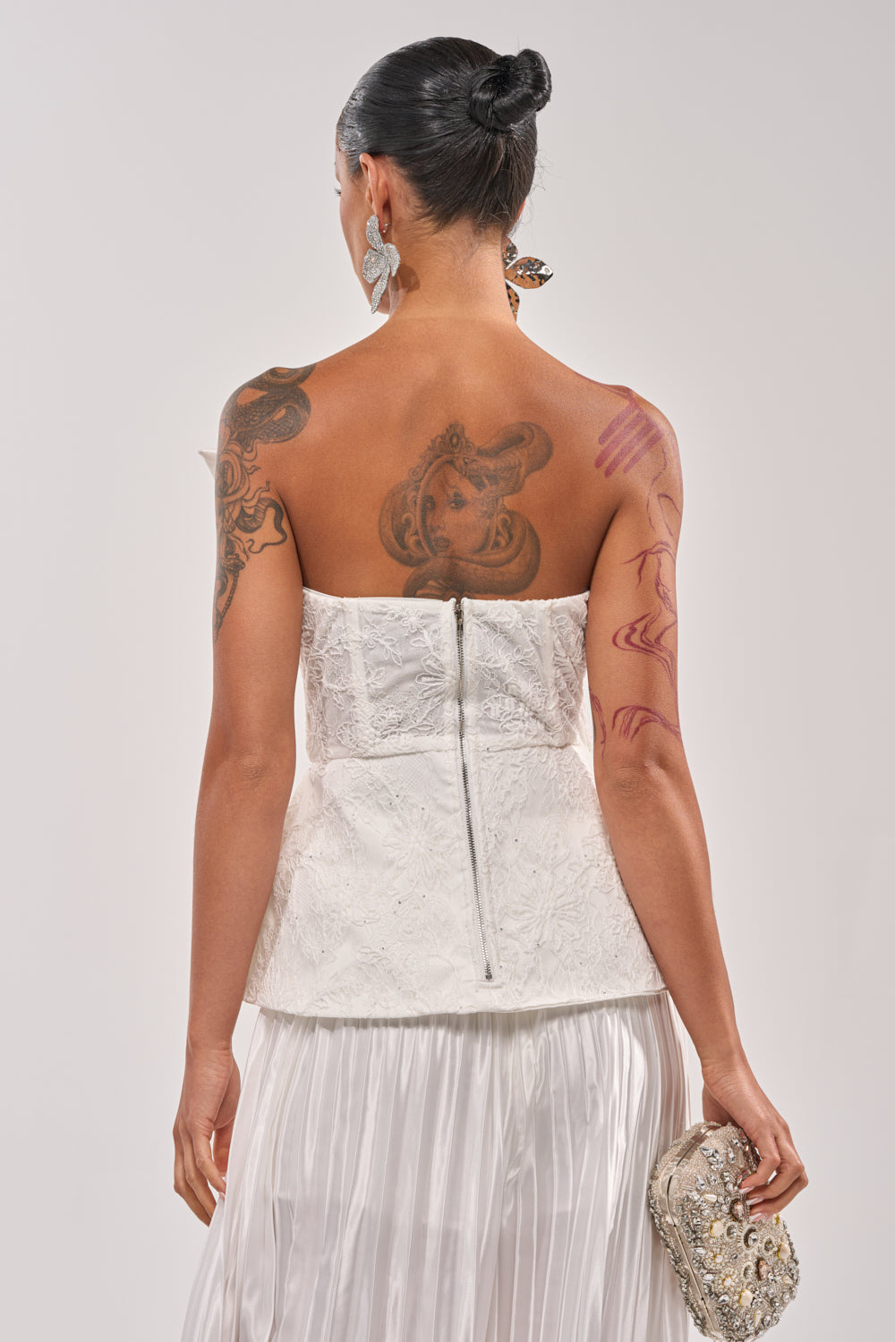 A woman with tattoos wears the CALLED OUT LACE CORSET and a pleated skirt, facing away while holding a jeweled clutch. Her sleek bun and bold silver earrings complete the look. 3
