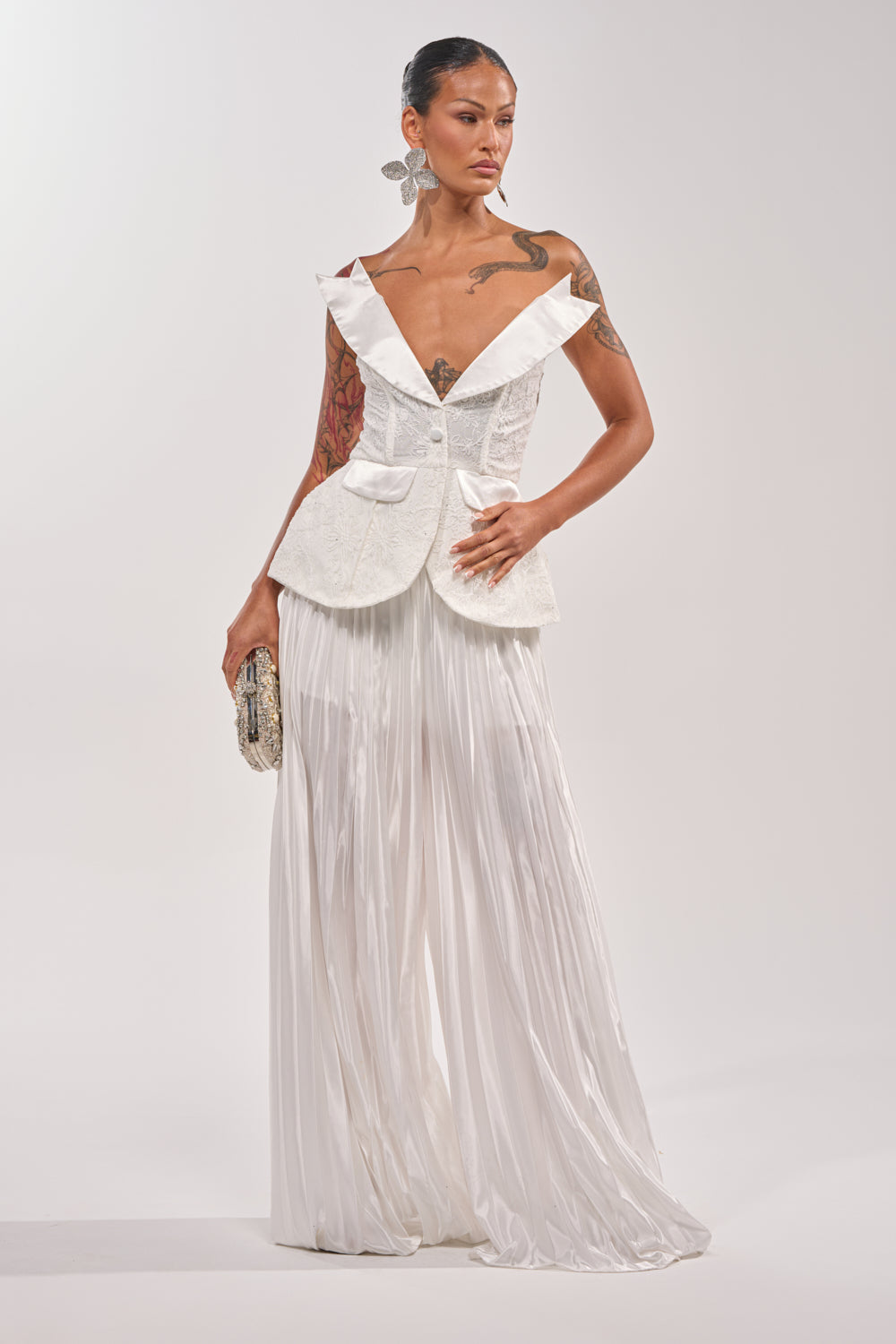 A woman exudes confidence in the CALLED OUT LACE CORSET, featuring a structured off-shoulder design, large lapels, and bow accents, paired with pleated pants. She completes the look with a clutch, tattoos, and statement earrings. 5