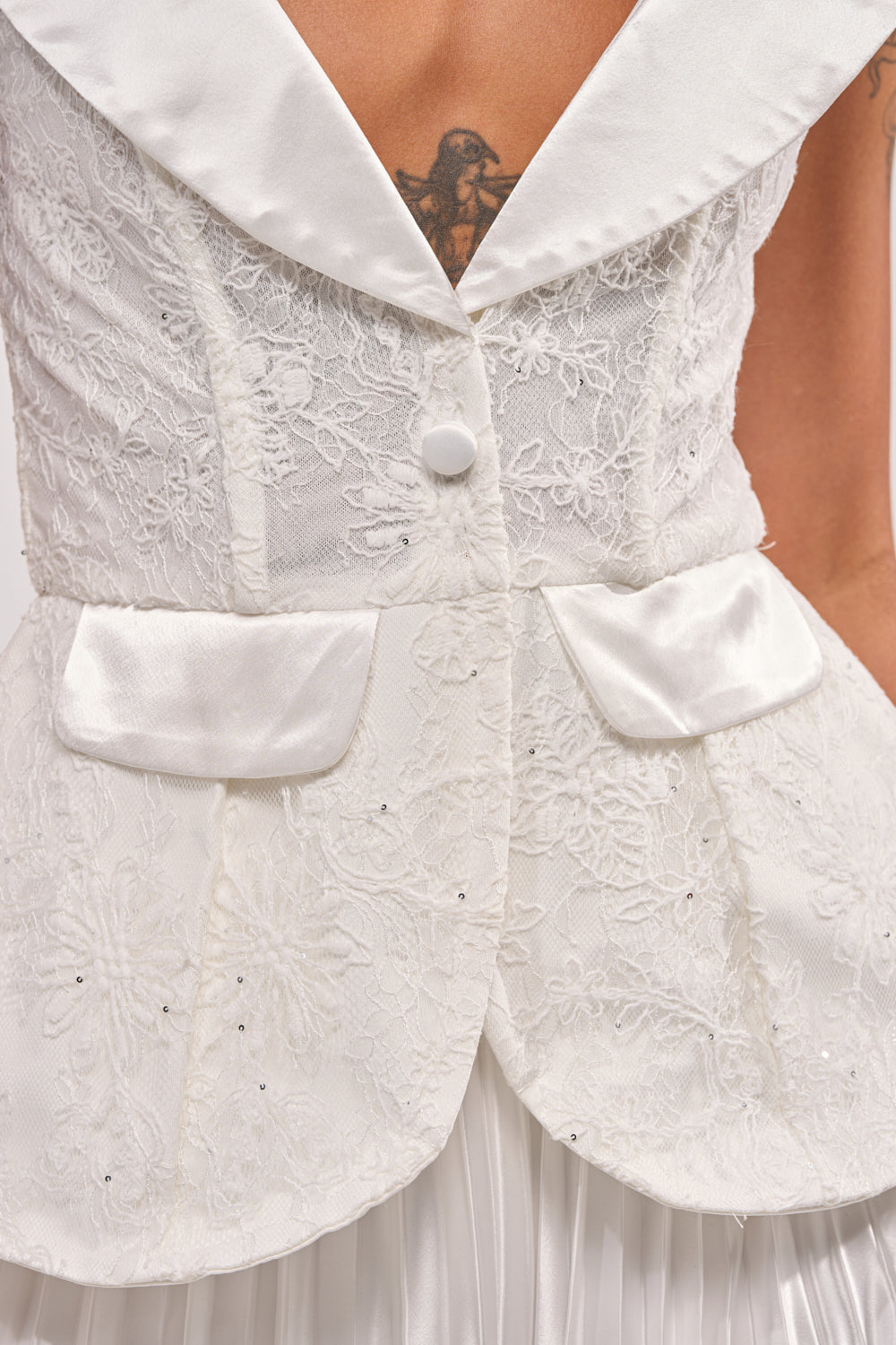 A close-up of the CALLED OUT LACE CORSET, a white lace top with satin lapels, pockets, and single button closure, styled over a pleated white skirt. A bird tattoo appears on the wearer’s chest. 6