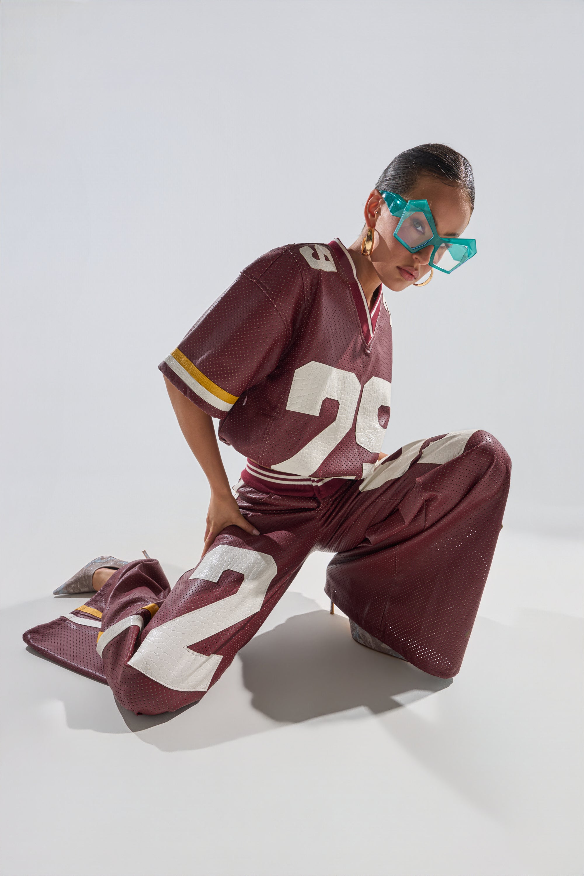 A person poses on one knee in a maroon football jersey and the LUCKY 29 TROUSER—sporty, perforated faux leather wide-leg pants with bold white numbers—plus oversized blue geometric glasses, against a plain light background. 2