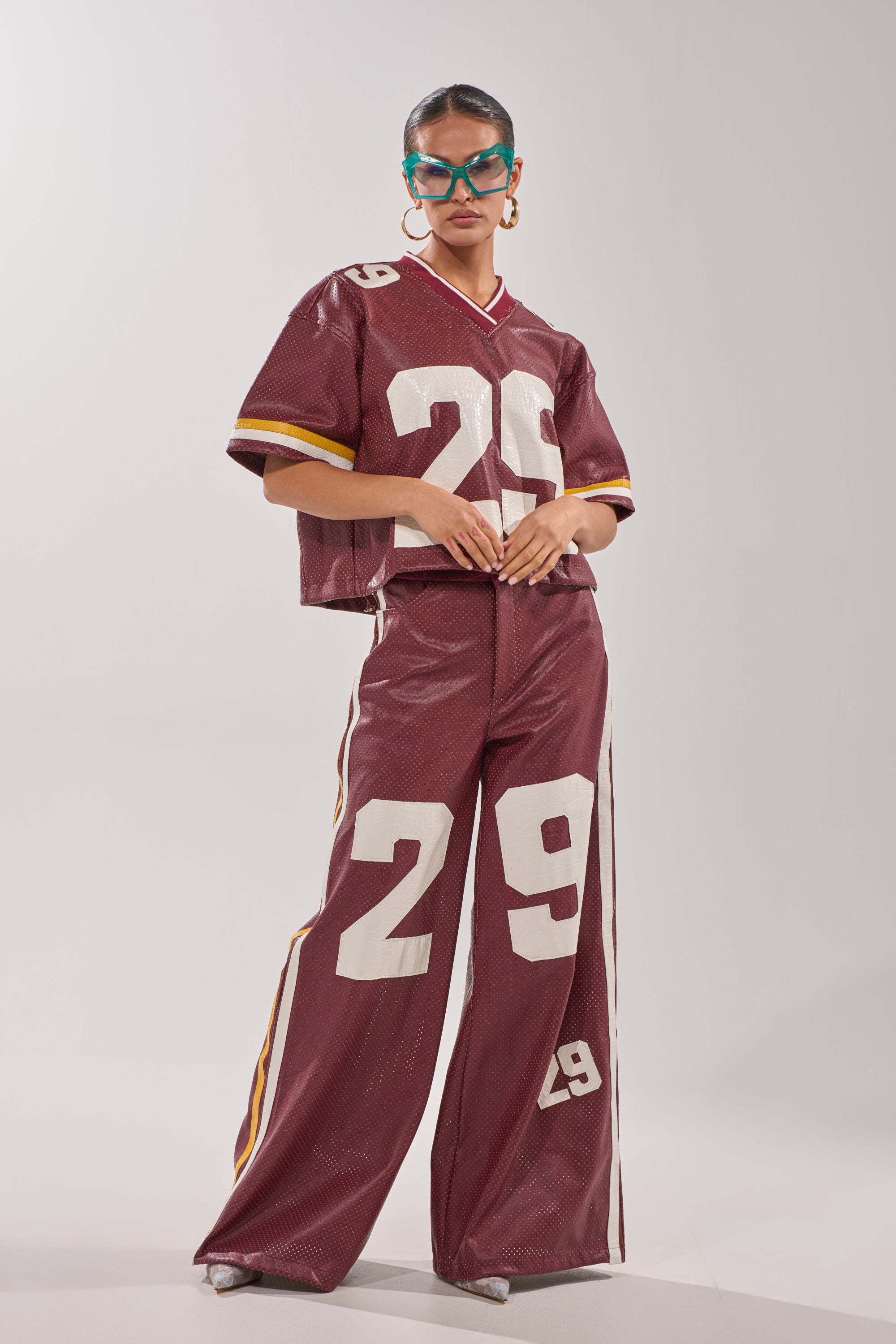A woman models the LUCKY 29 TROUSER in maroon with a perforated sporty design and the number 29 on both top and pants, styled with blue sunglasses and hoop earrings against a plain light background. 10