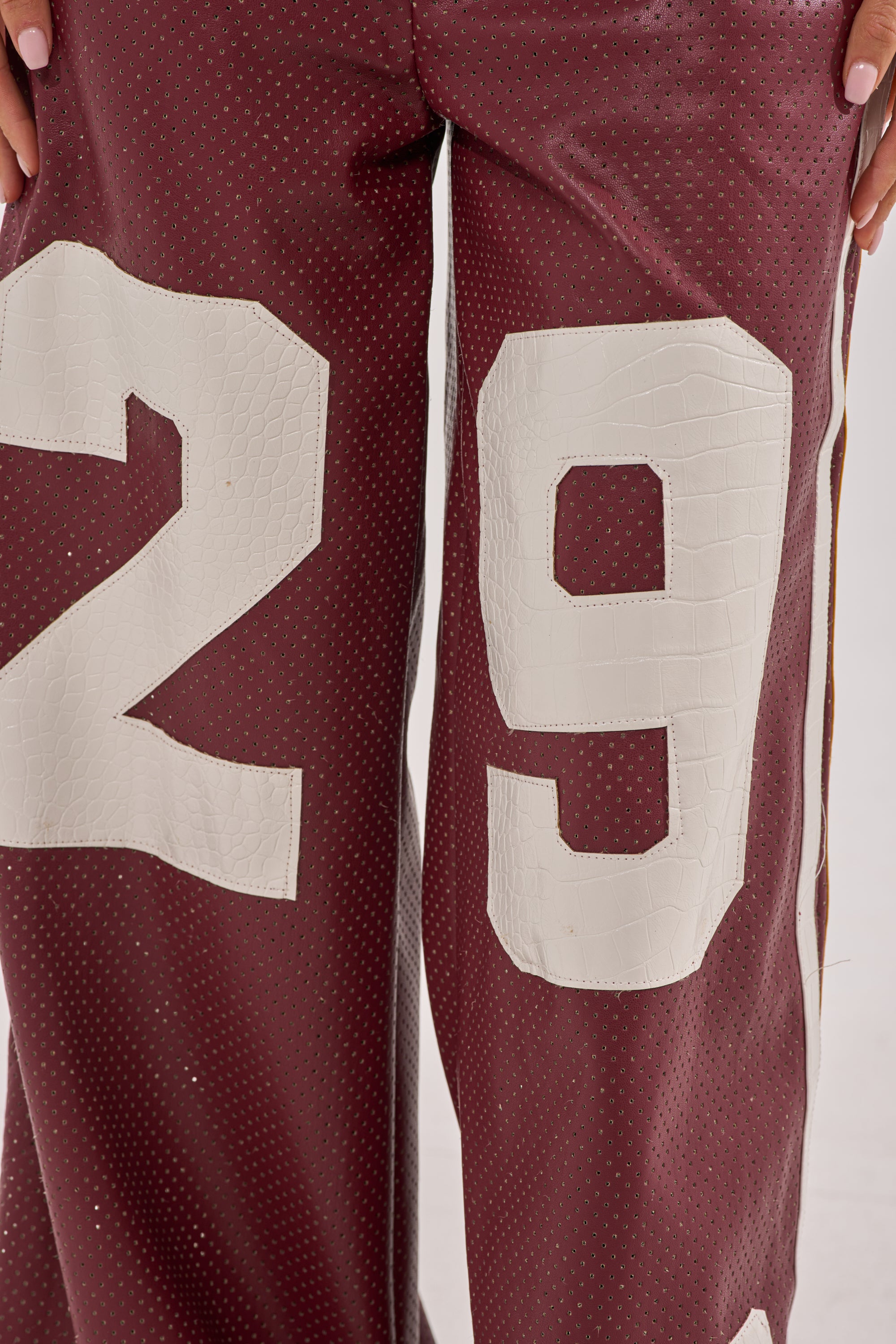 Close-up of a person wearing LUCKY 29 TROUSER—sporty burgundy pants with a perforated design and bold white "29" on the legs. The individual's hands are partially visible, set against a plain light background. 11