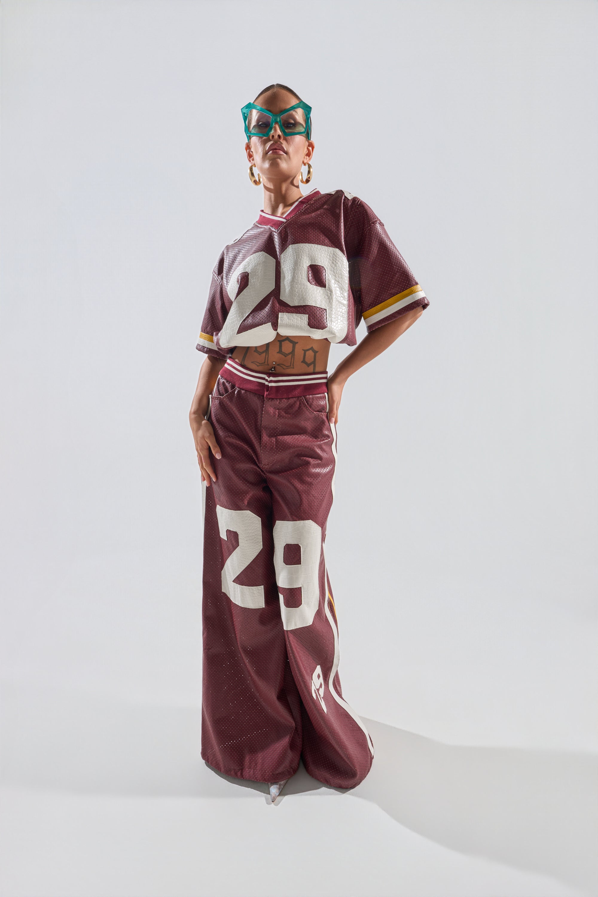 A person in teal sunglasses and hoop earrings poses confidently in the LUCKY 29 TROUSER set—a sporty burgundy outfit with perforated detail, featuring the number 29 in white on both the cropped top and wide-leg pants. 3