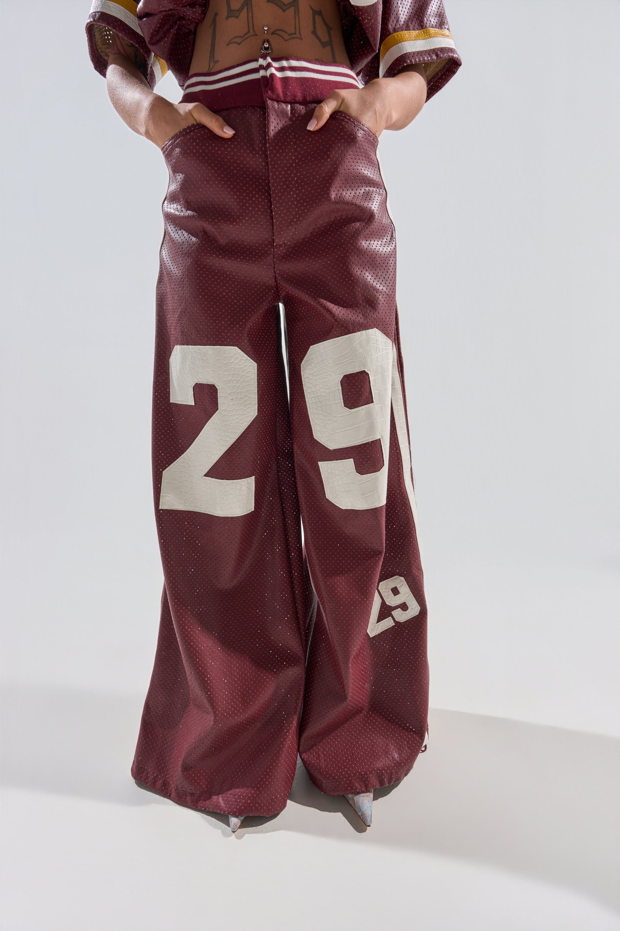 Model wearing the LUCKY 29 TROUSER—baggy burgundy faux leather wide-leg pants with bold white "29" on each leg, hands in pockets, posed against a plain light background. Face and upper body are not shown. 1