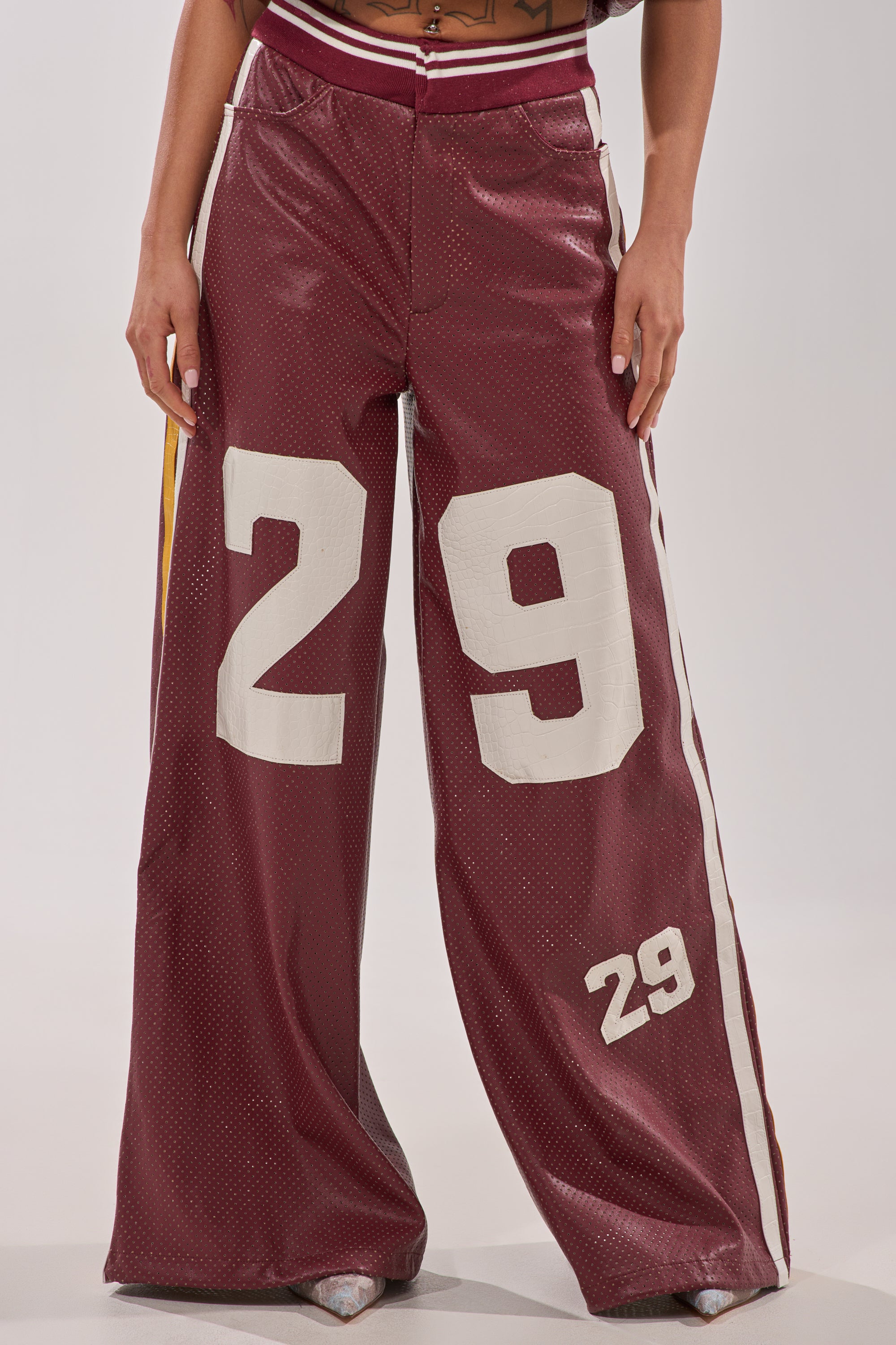 A person wearing the LUCKY 29 TROUSER: burgundy faux leather wide-leg pants featuring shiny croc-embossed side stripes and bold white "29" numbers on the left thigh and lower right leg. 0