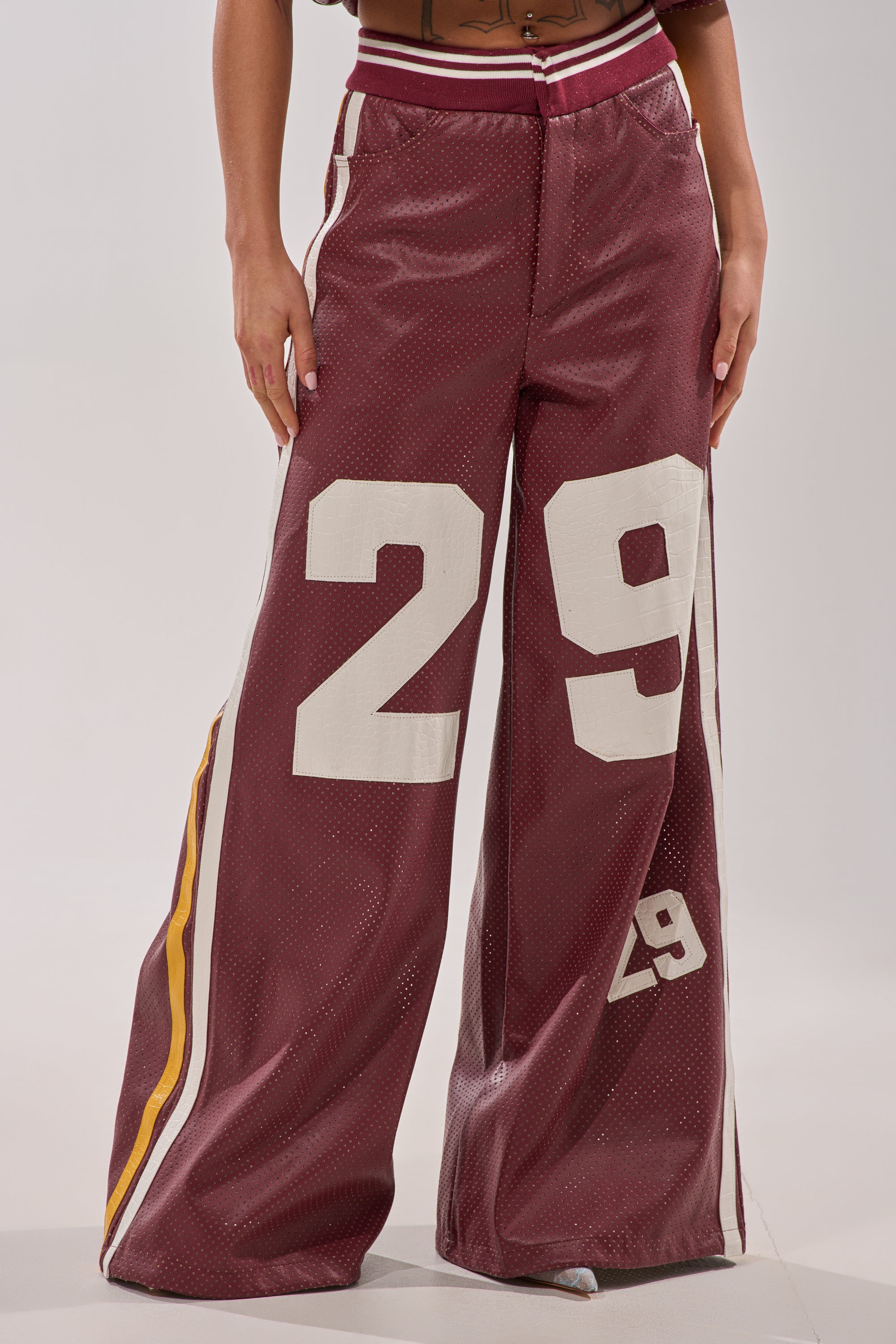A person wears the LUCKY 29 TROUSER: wide-leg burgundy pants with a bold white "29" on one leg, a smaller "29" on the other, and yellow and white side stripes. The image is cropped at the waist and ankles. 5