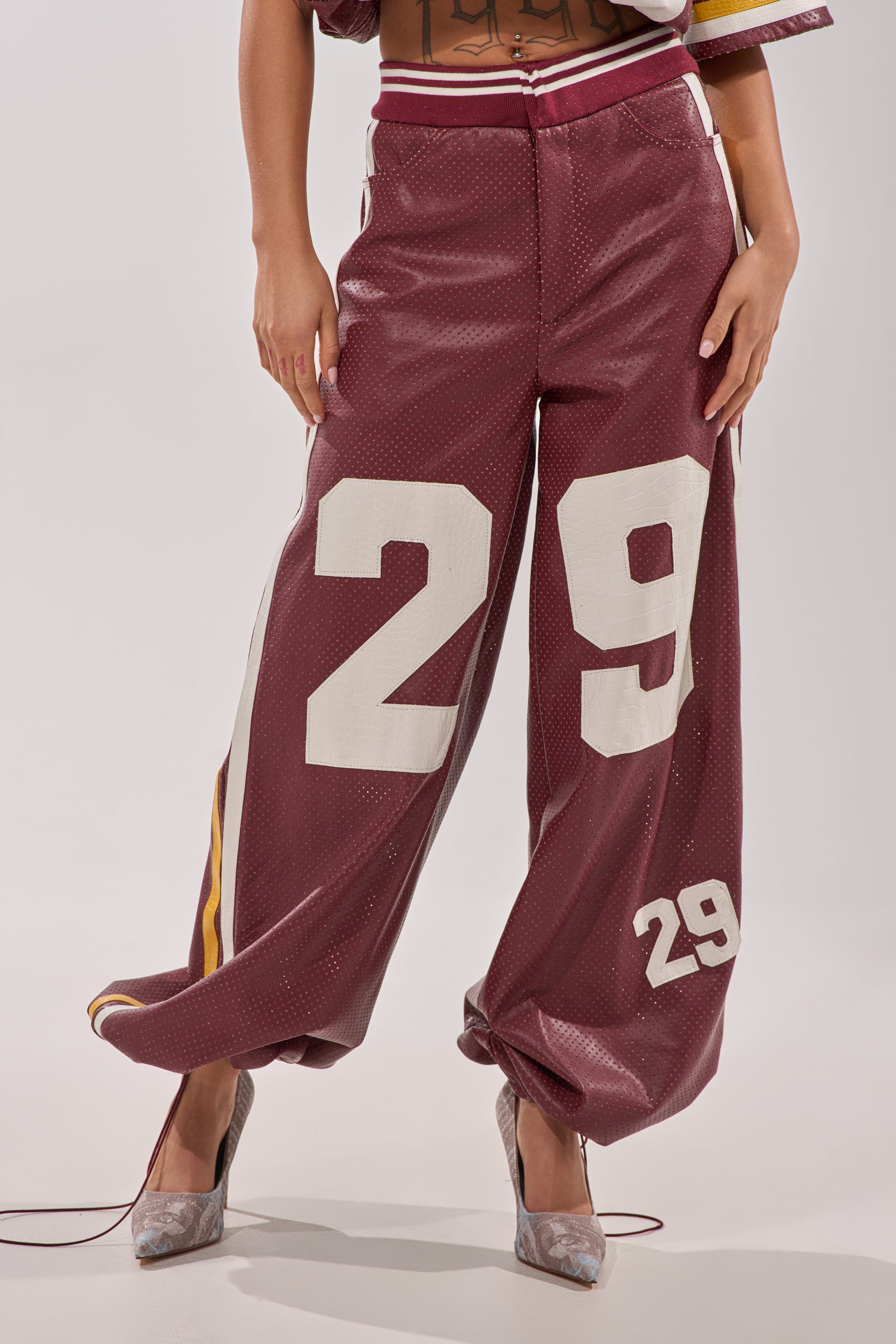 A person models the LUCKY 29 TROUSER—loose burgundy pants with sporty perforations and bold white "29" graphics—teamed with silver high heels. Only their lower half appears against a plain background. 6