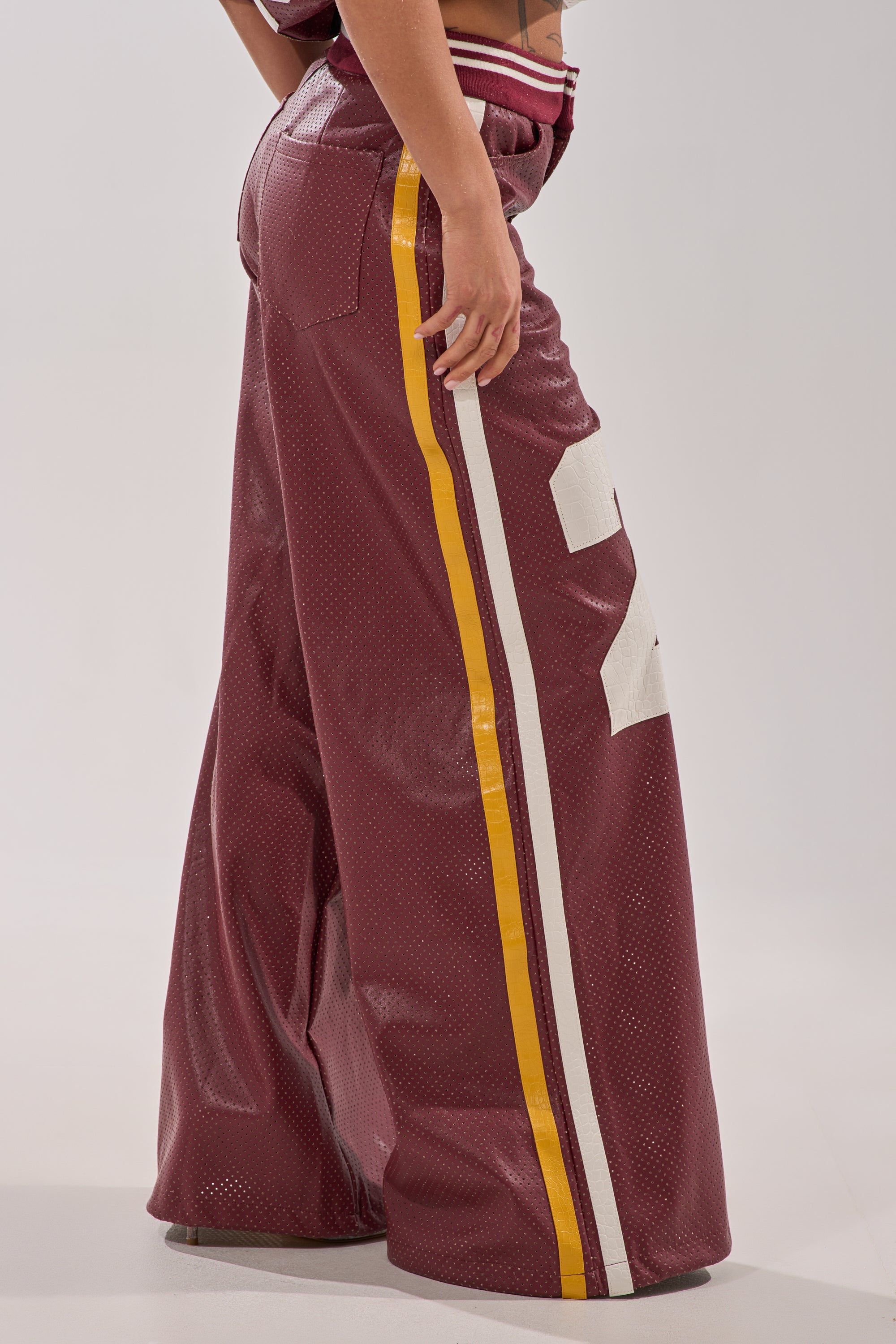 A person wears the LUCKY 29 TROUSER—burgundy wide-leg pants with yellow and white side stripes and bold white numbers—standing against a plain background. 7