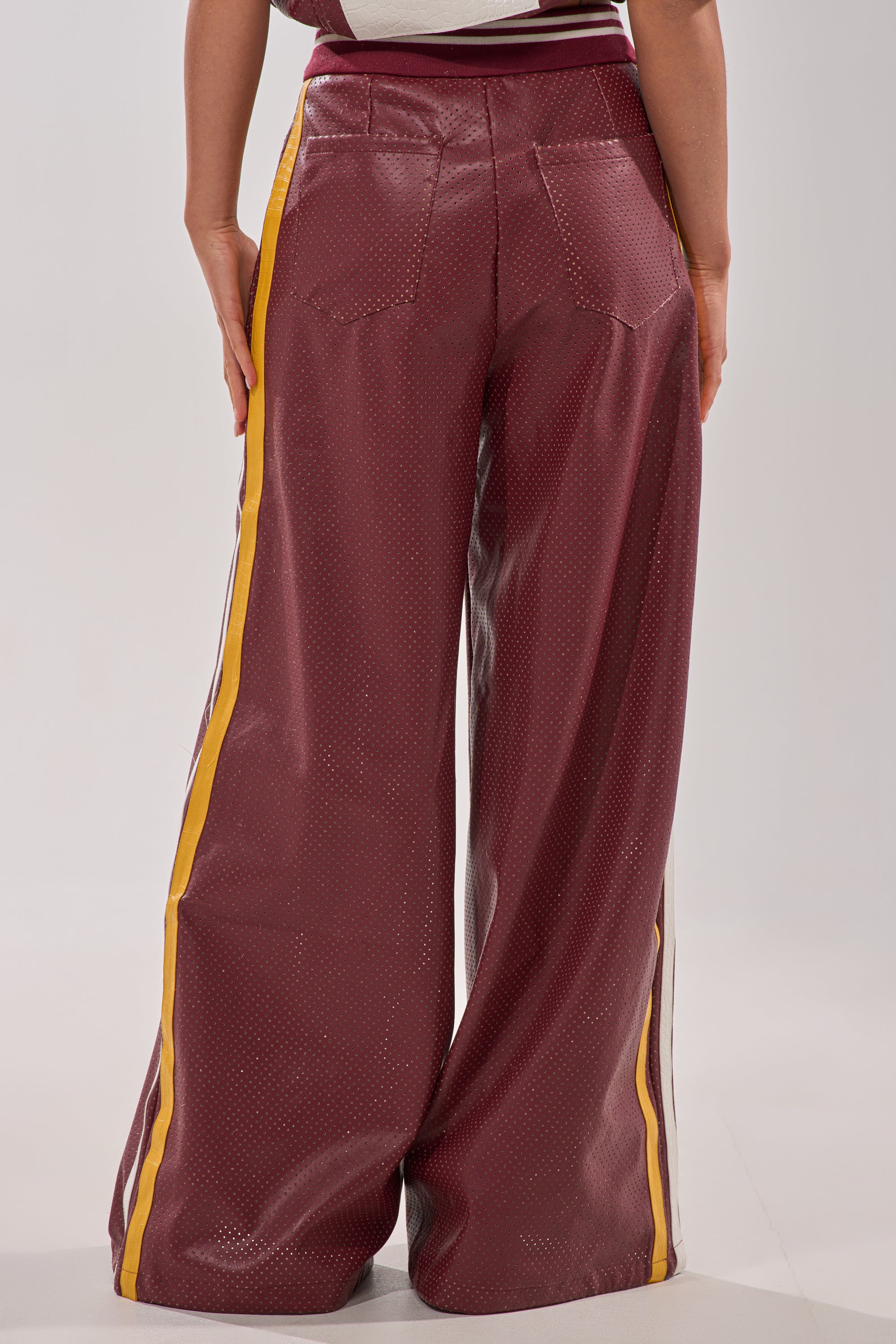 Someone is seen from behind, wearing the LUCKY 29 TROUSER—burgundy faux leather wide-leg pants with yellow and white side stripes—standing against a neutral background. 8