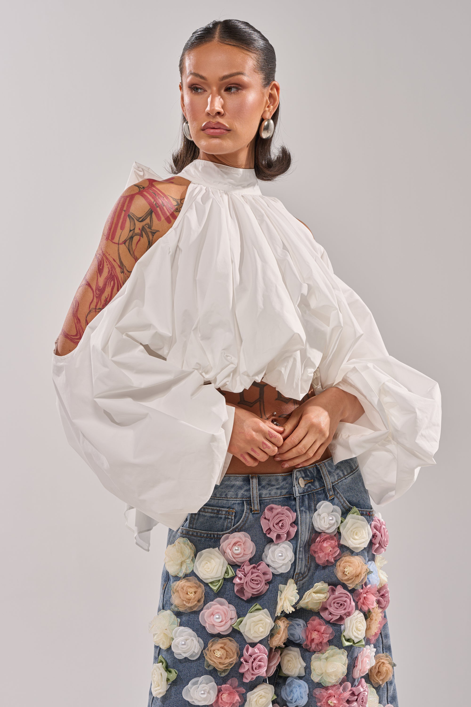 A woman with long dark hair and a visible arm tattoo wears the ALEXIA MOCK NECK BLOUSE IN WHITE, featuring voluminous bubble hem and open-shoulder sleeves, styled with blue jeans adorned with colorful 3D fabric flowers against a plain background. 2
