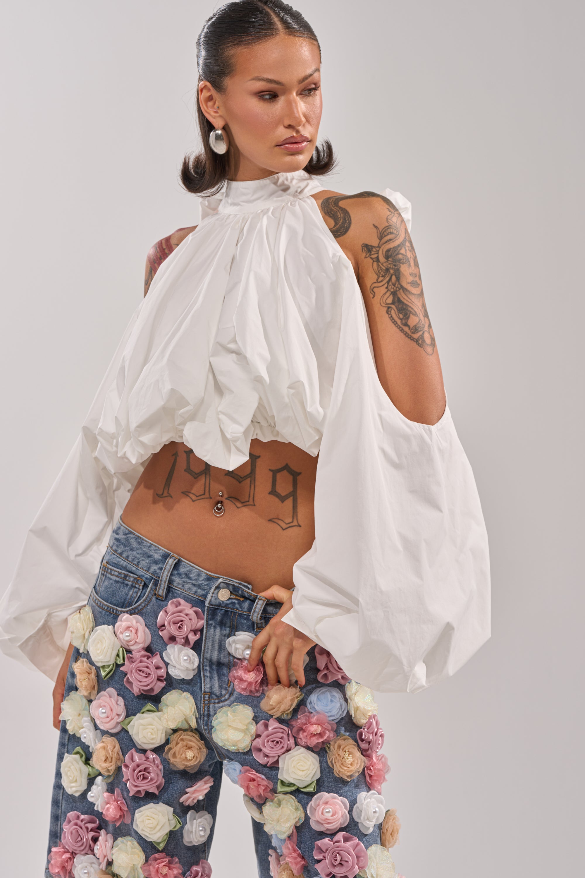 A woman with tattoos wears the ALEXIA MOCK NECK BLOUSE IN WHITE—billowy with open-shoulder sleeves—and high-waisted jeans adorned with colorful fabric flowers. Her hair is slicked back as she poses, looking to the side. 3