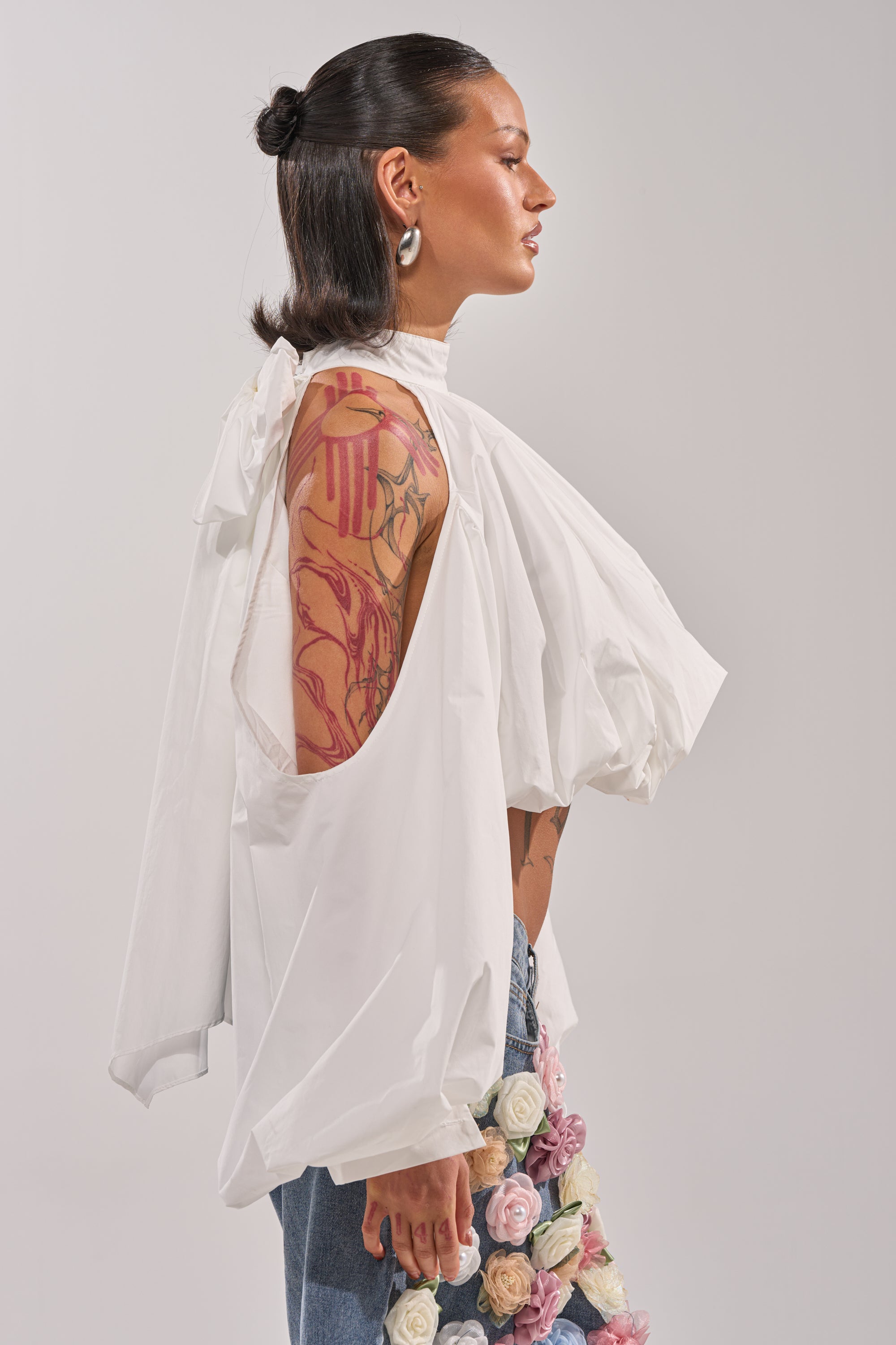 A person with slicked-back dark hair wears the ALEXIA MOCK NECK BLOUSE IN WHITE with open sleeves and blue jeans featuring fabric flowers. In profile, their visible arm shows off colorful floral tattoos. 4