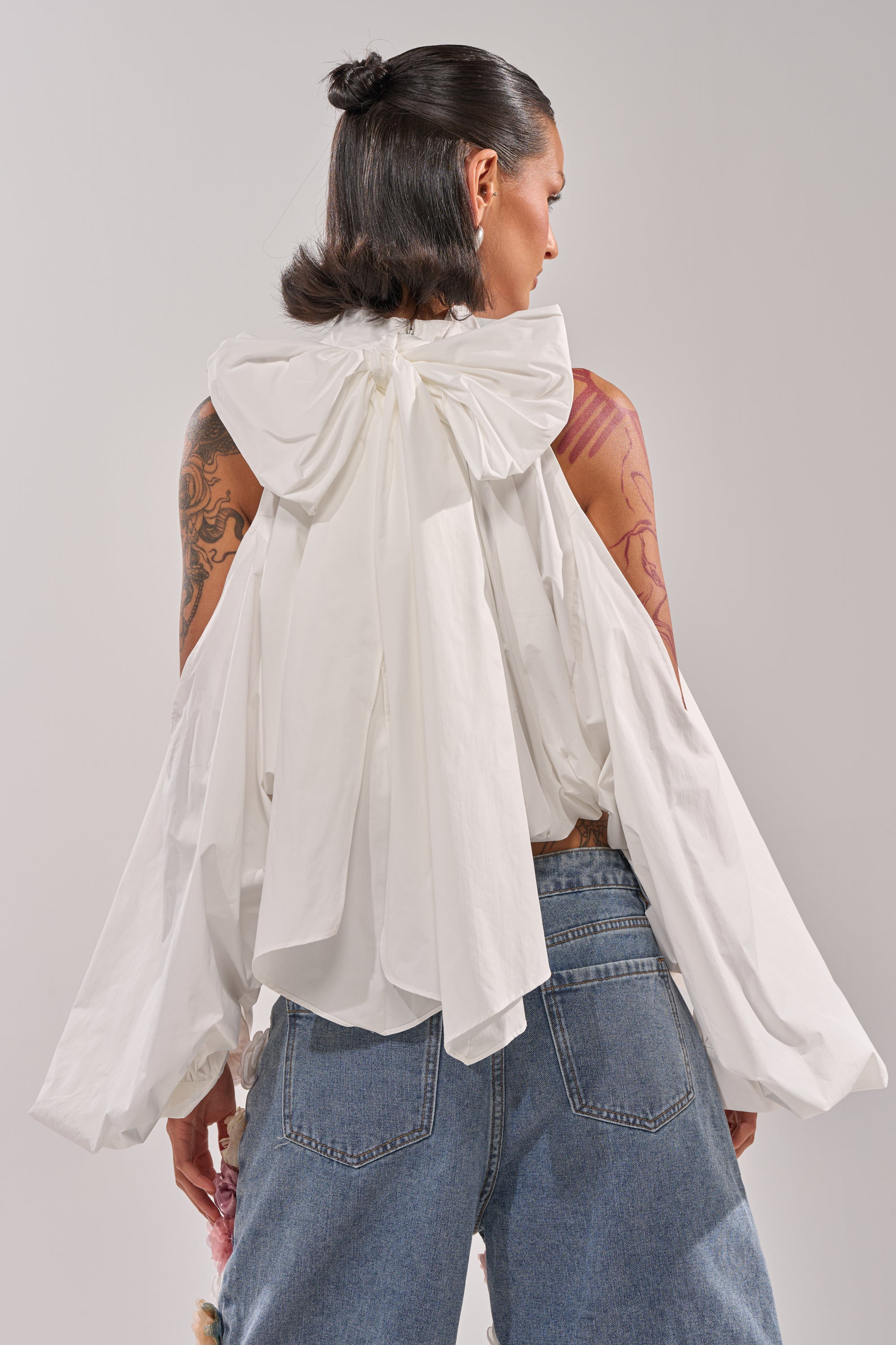 A person with dark hair in a half-up style is seen from behind, wearing the ALEXIA MOCK NECK BLOUSE IN WHITE, featuring a large bow at the collar and puffy sleeves, paired with blue jeans and revealing tattoos on their arms. 5