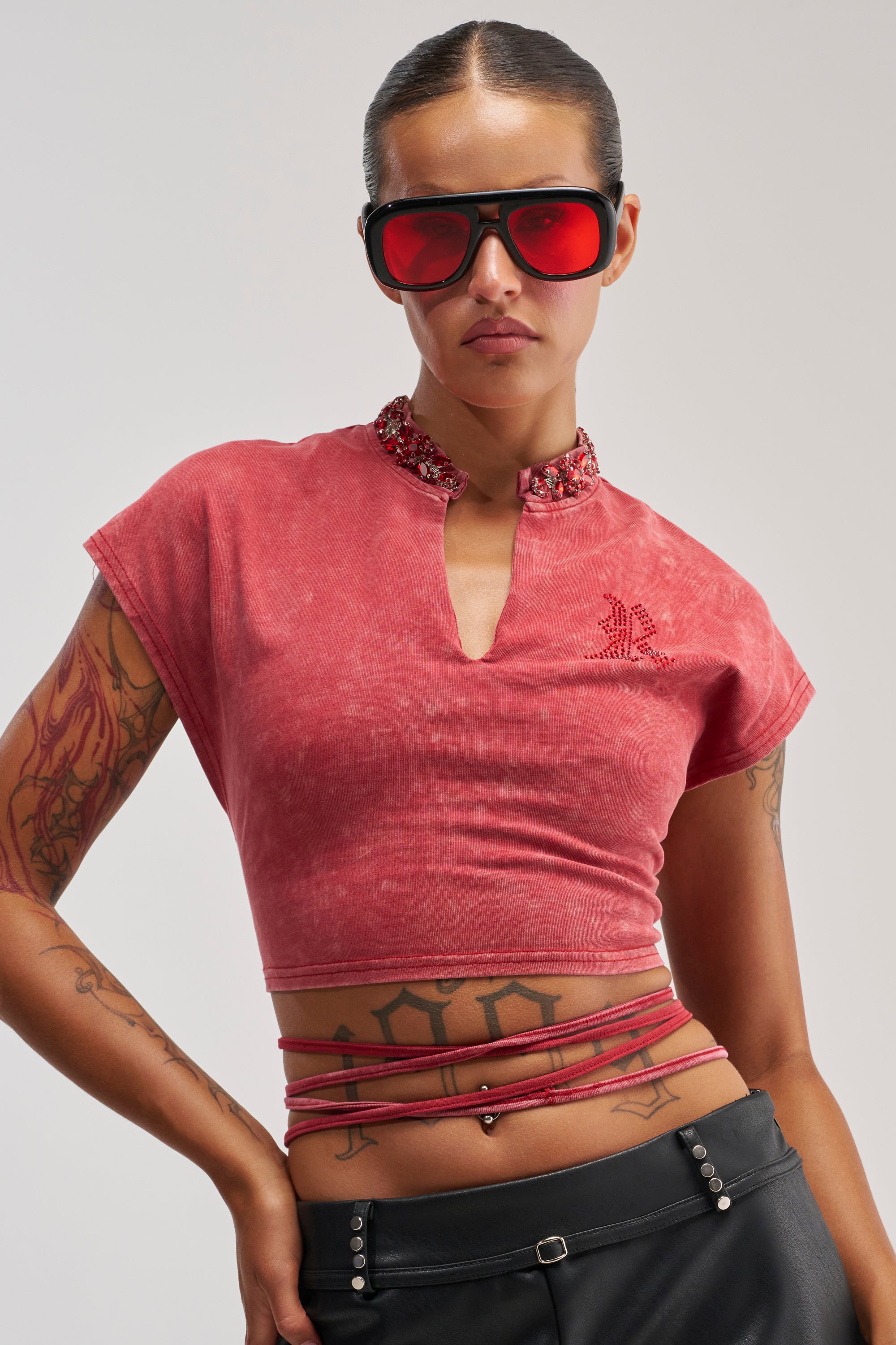 A woman stands confidently against a plain background wearing red sunglasses and the CHERRY ON TOP CROPPED TEE, a streetwear top featuring a beaded rhinestone collar. Her bold festival look is completed with eye-catching string details at the waist, black pants, and her arm tattoos on display.