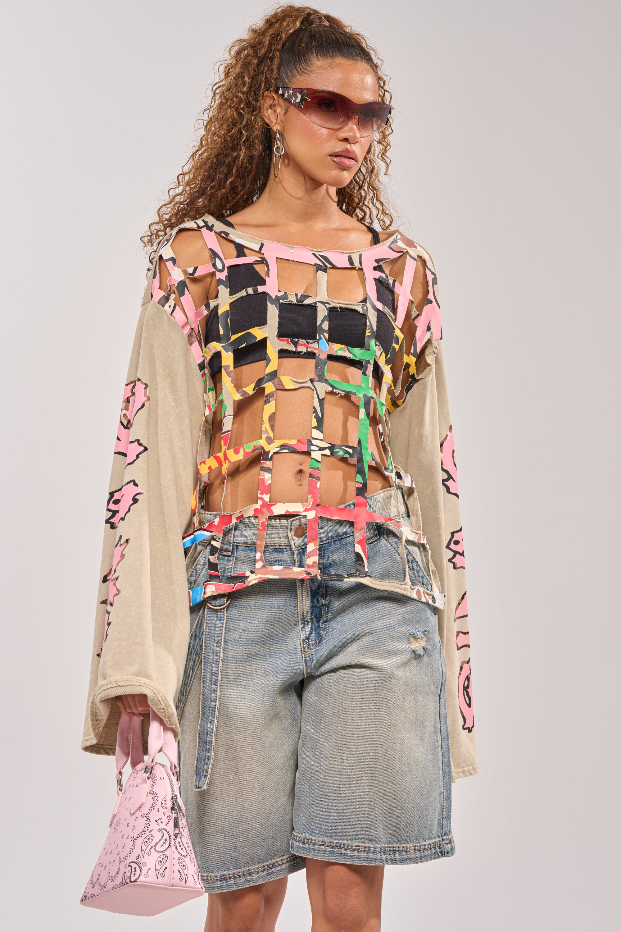 A woman with curly hair rocks sunglasses, a sheer grid-patterned top with colorful ribbons over a crop top, oversized denim shorts, and the CAGED UP LONG SLEEVE GRAPHIC TEE, holding a pink patterned handbag—streetwear layering perfection. 0