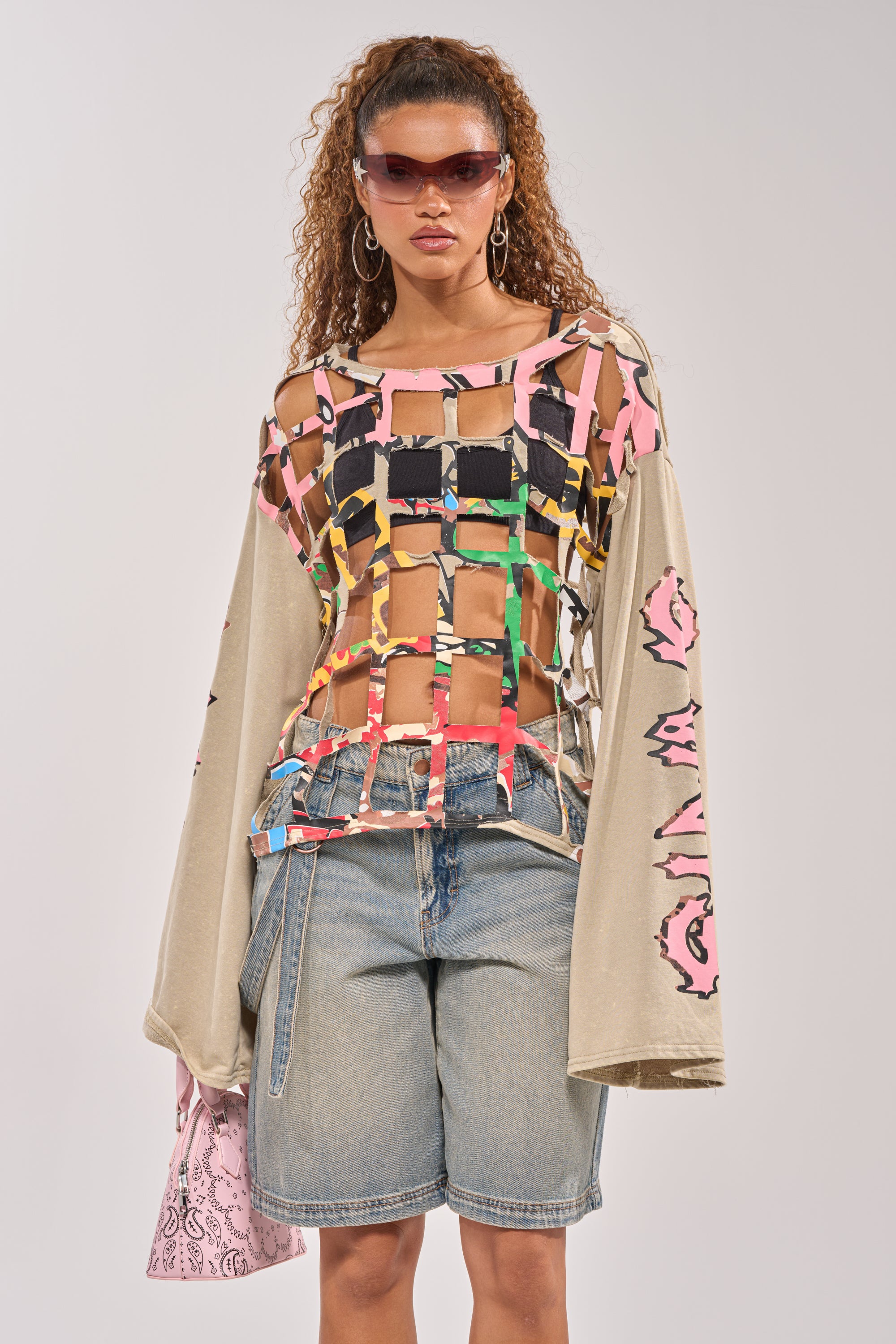 A woman with long curly hair and sunglasses wears hoop earrings, a CAGED UP LONG SLEEVE GRAPHIC TEE over a bandeau, wide-sleeved beige shirt with pink patterns, denim Bermuda shorts, and carries a pink patterned bag. 1