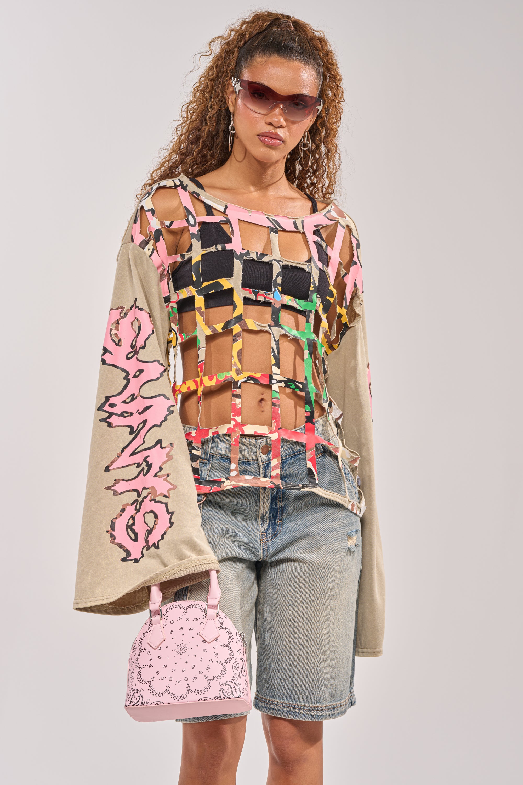 A woman with curly hair and a confident look stands against a plain background, wearing sunglasses, knee-length denim shorts, a pink handbag, and the CAGED UP LONG SLEEVE GRAPHIC TEE styled with colorful ribbons. 2