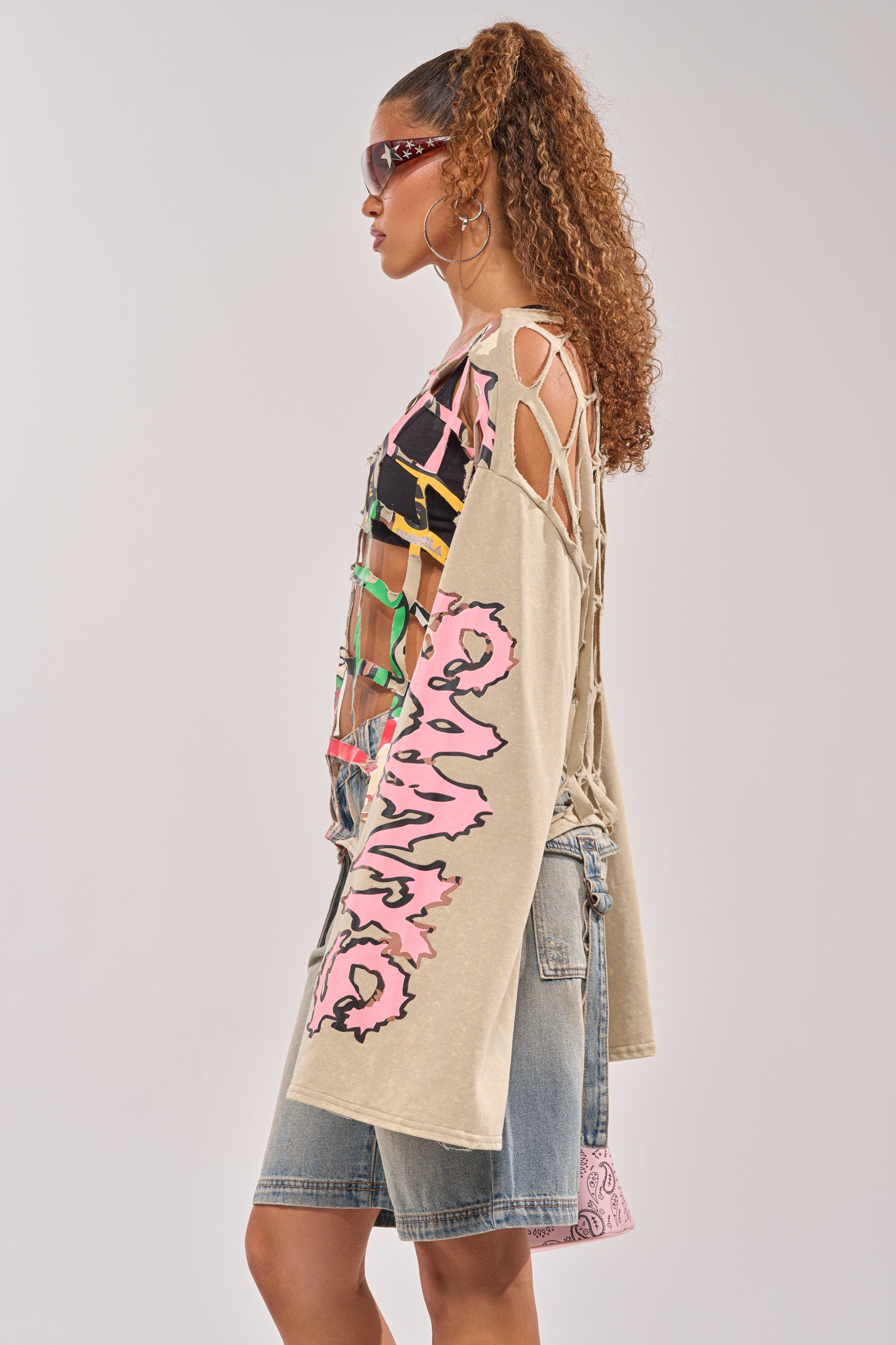 A woman with curly hair in a ponytail wears sunglasses, large hoop earrings, denim shorts, and the CAGED UP LONG SLEEVE GRAPHIC TEE with pink sleeve designs, standing in profile against a plain background. 3