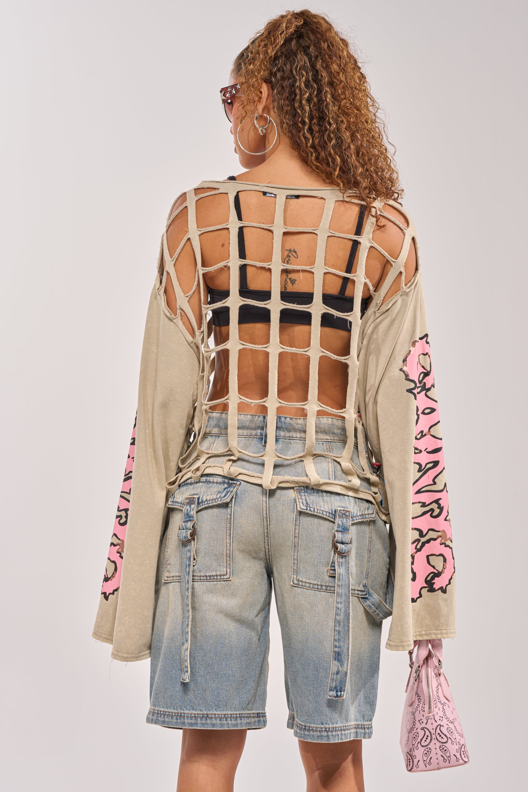 A person with curly hair and hoop earrings wears the CAGED UP LONG SLEEVE GRAPHIC TEE with a large open netted back, paired with denim shorts featuring side zippers, and holds a pink patterned bag. Pink accents decorate this streetwear look. 4