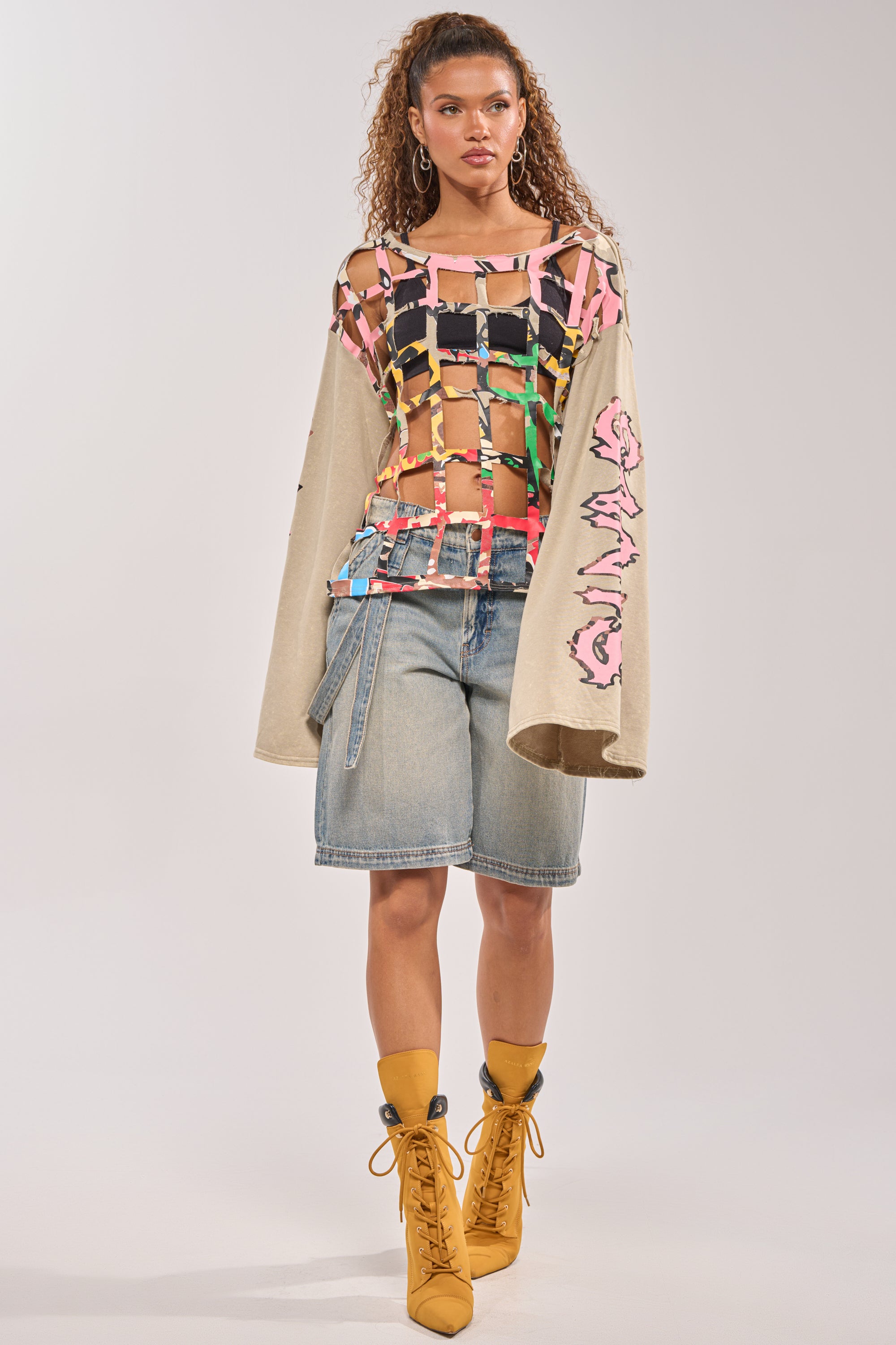 A woman wears a colorful patchwork mesh top over the CAGED UP LONG SLEEVE GRAPHIC TEE, paired with denim knee-length shorts and mustard yellow lace-up boots, standing against a plain background. 5