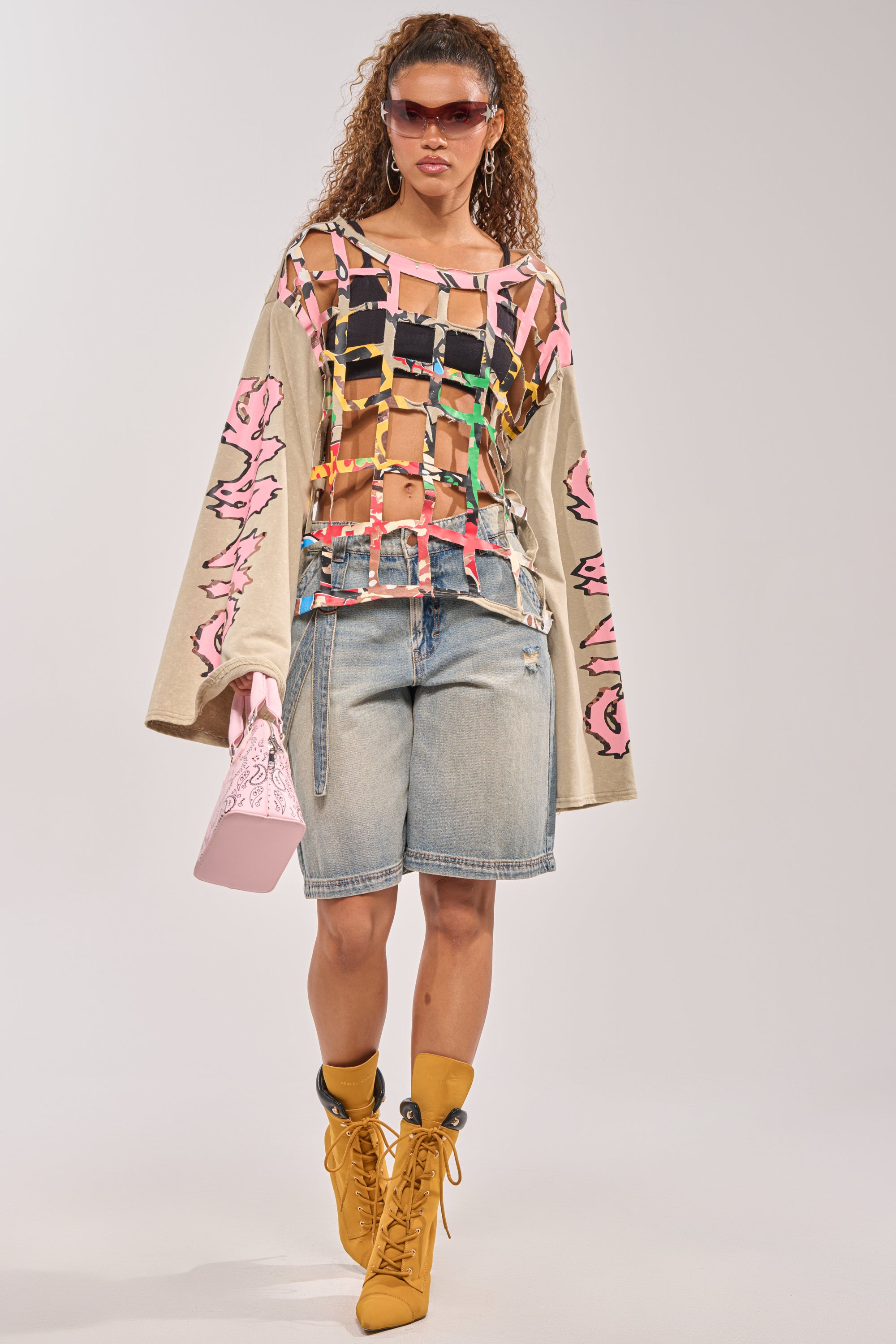 A woman in sunglasses, knee-length denim shorts, yellow lace-up boots, and a light pink handbag struts confidently in a CAGED UP LONG SLEEVE GRAPHIC TEE with pink and beige sleeves against a plain backdrop. 6