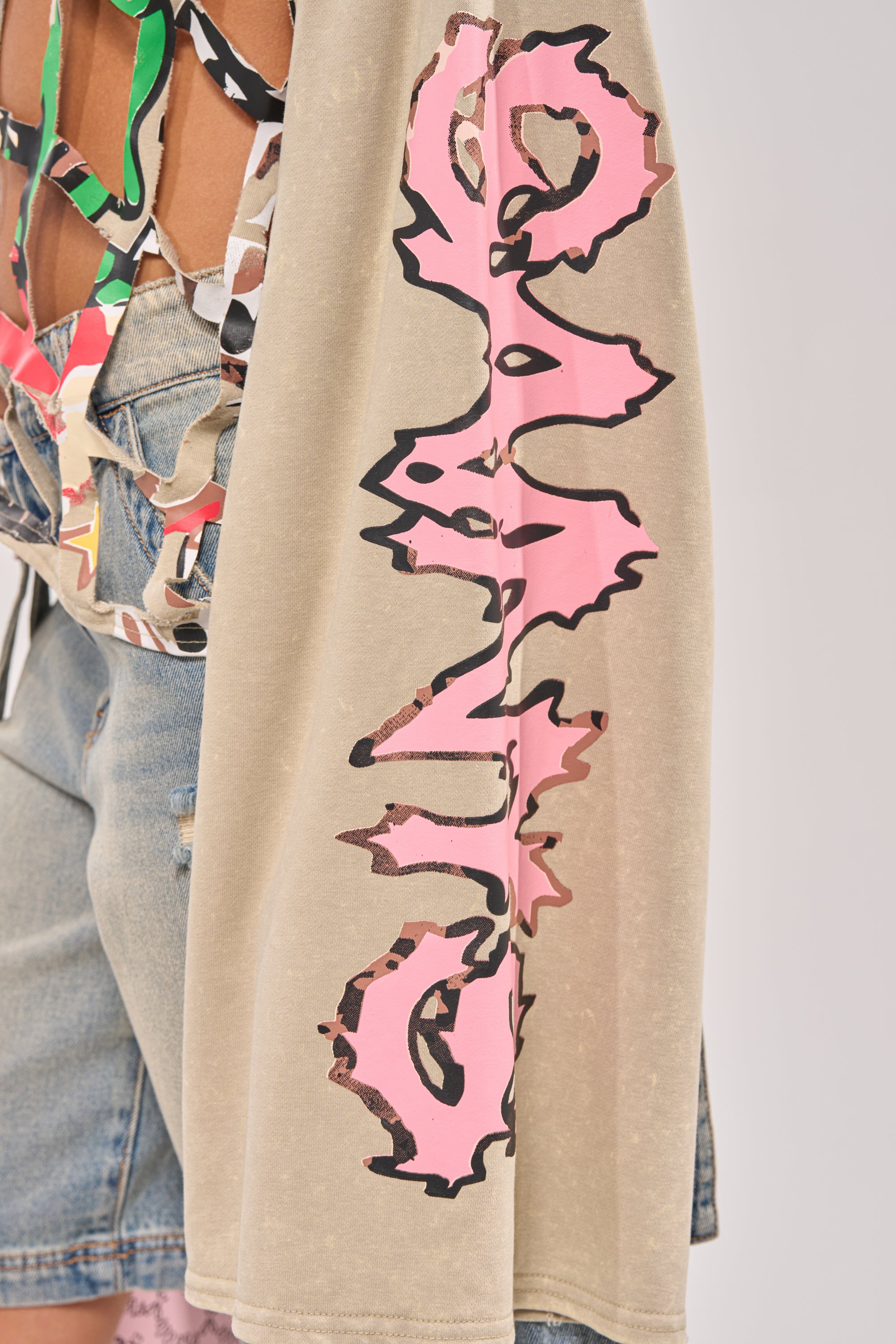 A close-up of a person wearing the CAGED UP LONG SLEEVE GRAPHIC TEE, a beige distressed streetwear top with pink and black sleeve graphics, styled with denim shorts and a colorful strappy top. 8