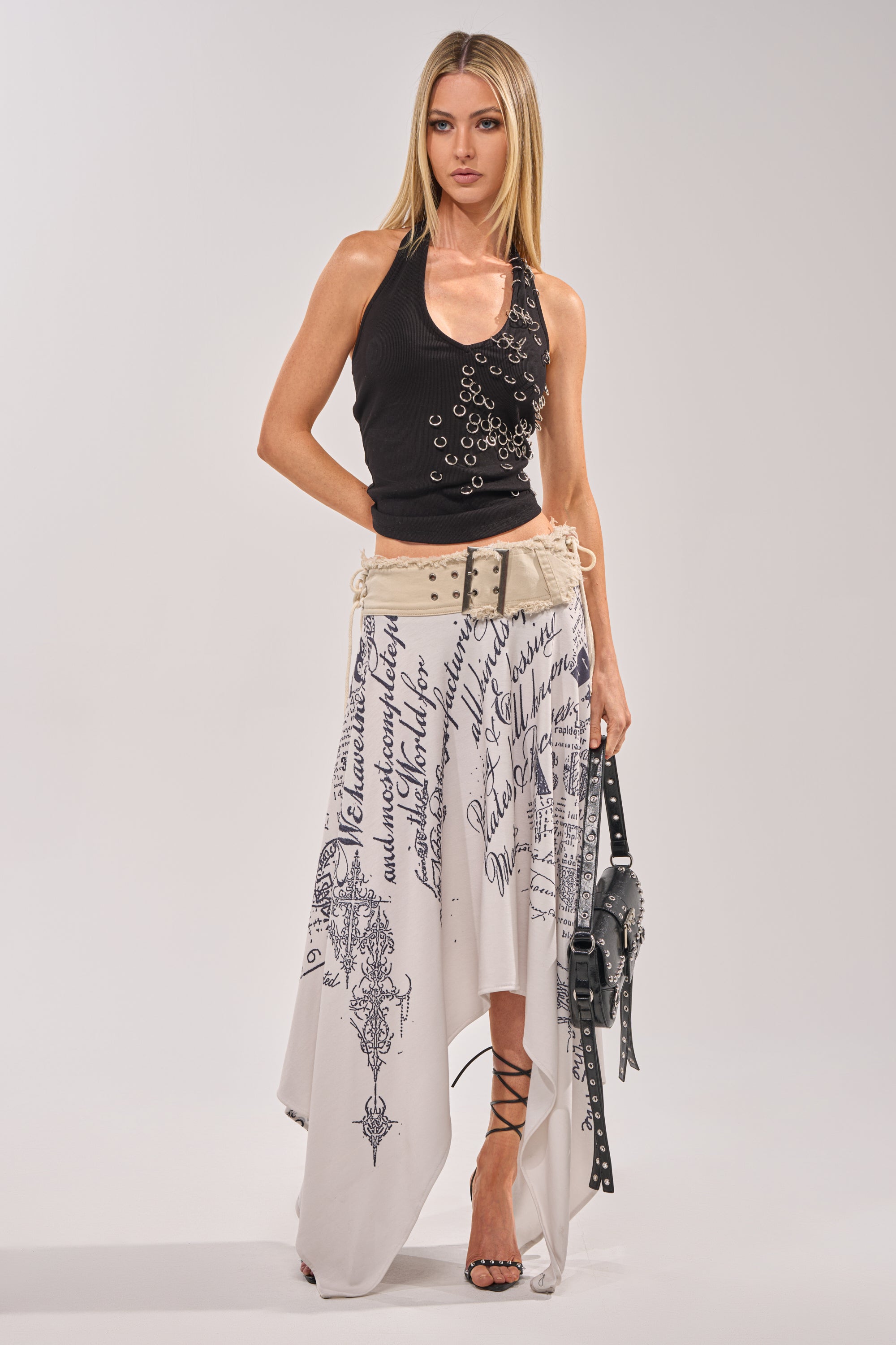 A woman stands against a plain background wearing the RING RING RING EMBELLISHED TANK IN BLACK, a long white asymmetrical skirt with black script and patterns, black strappy heels, and holding a black handbag. 5