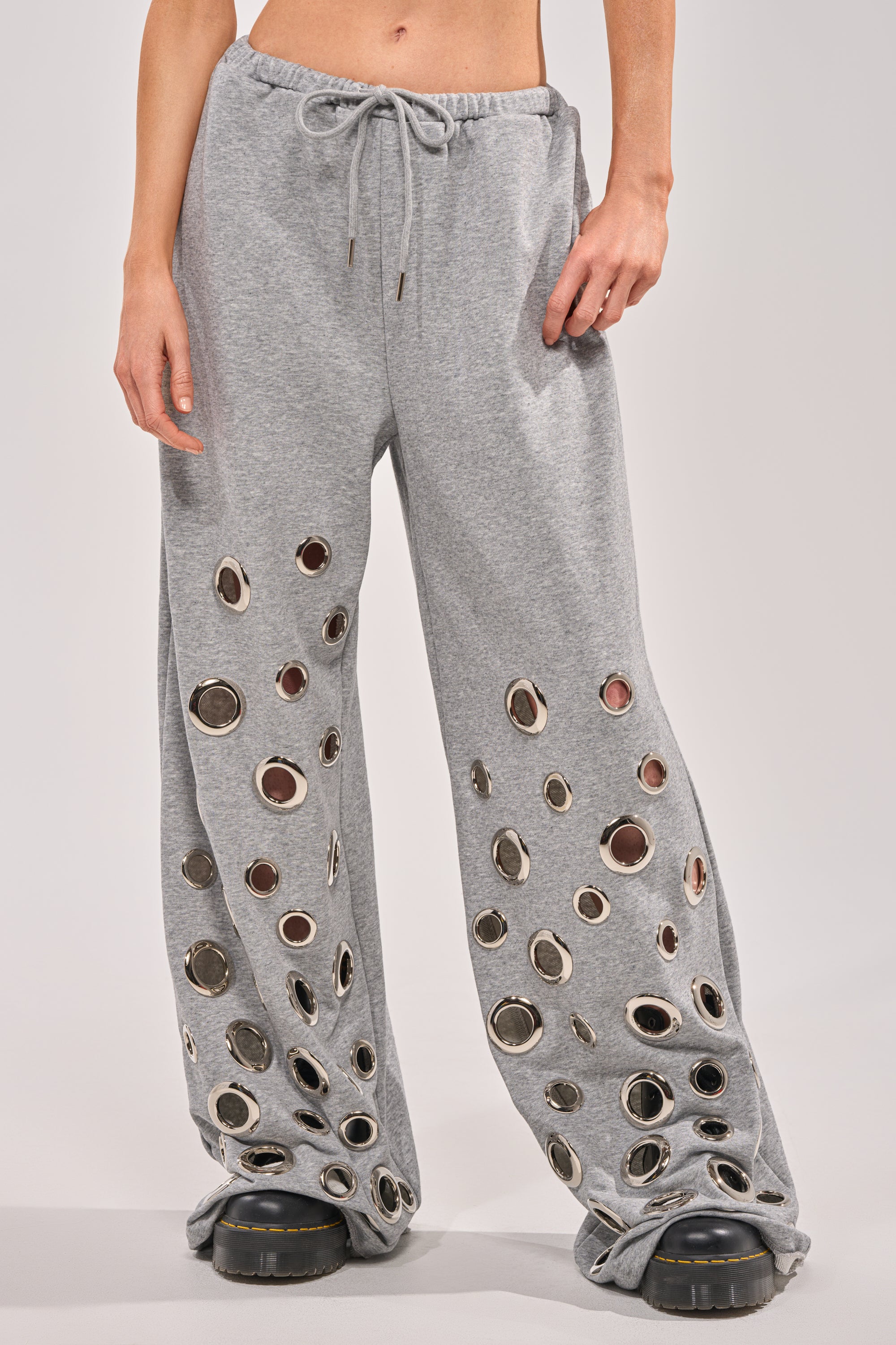 A person is seen from the waist down, wearing the CHECK IT OUT GROMMET JOGGER—grey wide-leg sweats with bold grommet hardware—paired with chunky black shoes against a plain background. 1