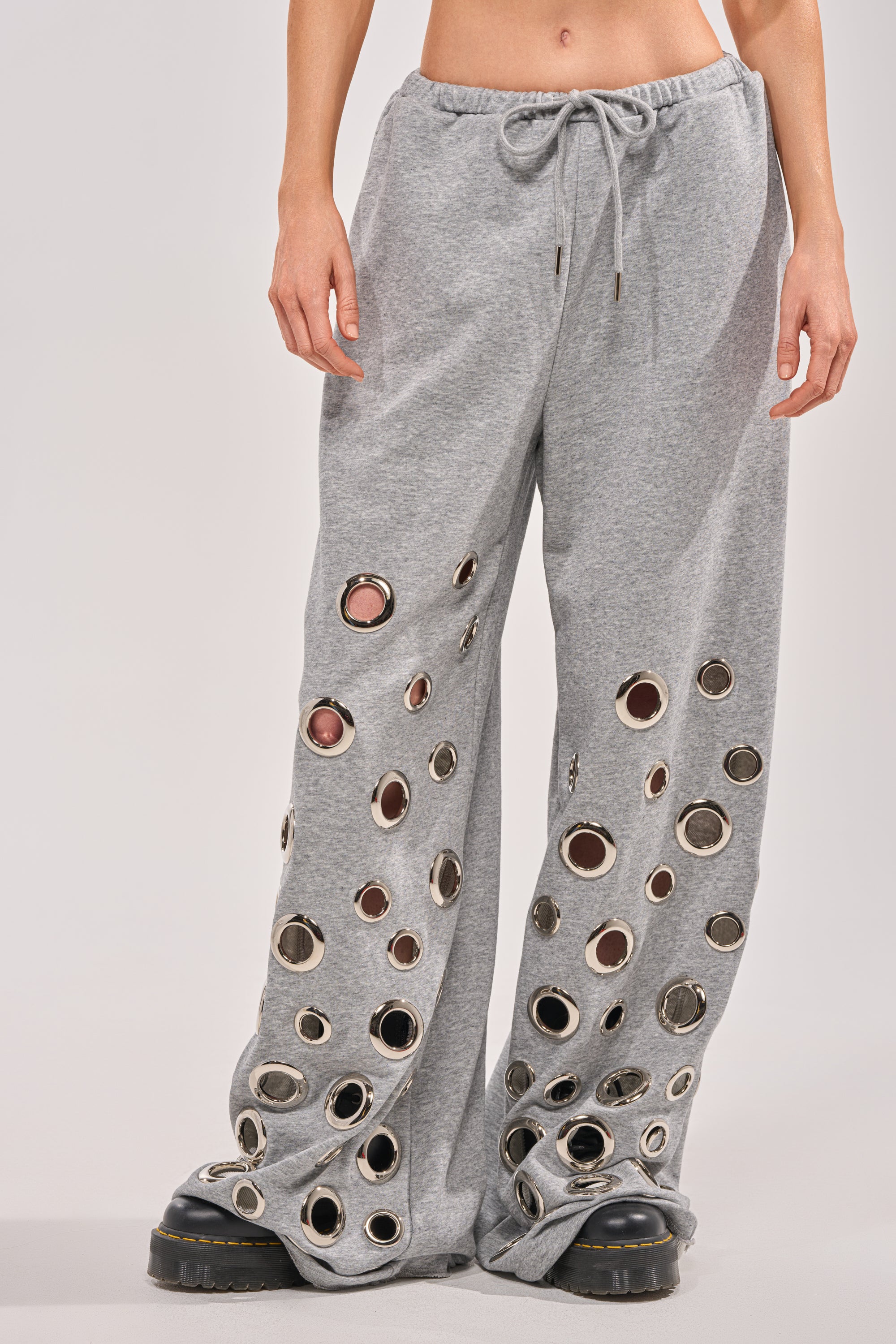 Wearing the CHECK IT OUT GROMMET JOGGER—loose grey wide-leg pants with bold grommet hardware and a drawstring waist—and black platform shoes, someone stands against a plain light background. 2
