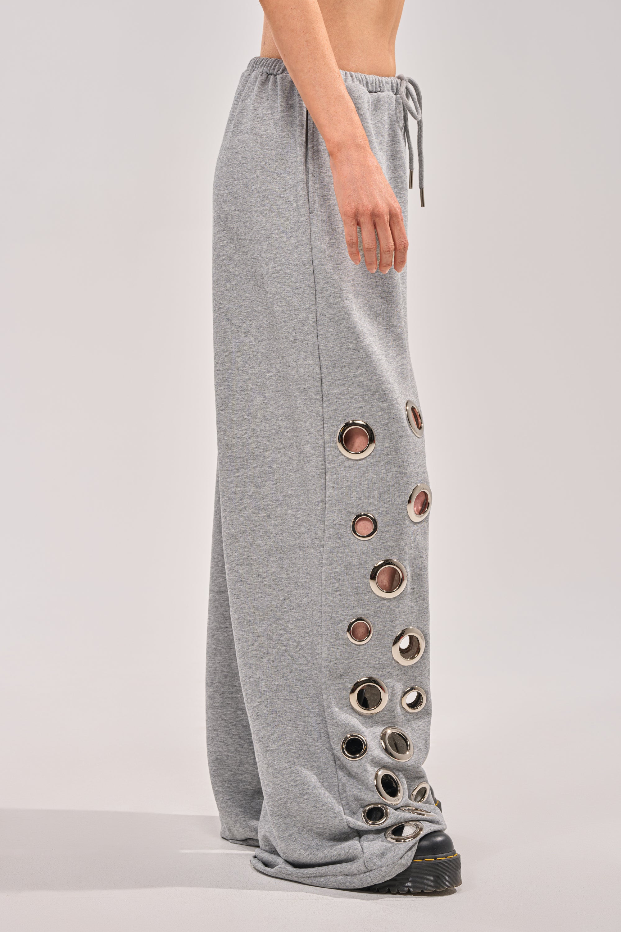 Someone wears the CHECK IT OUT GROMMET JOGGER, high-rise pants with bold silver grommet detailing on one leg, paired with chunky black shoes, standing against a plain background. 3