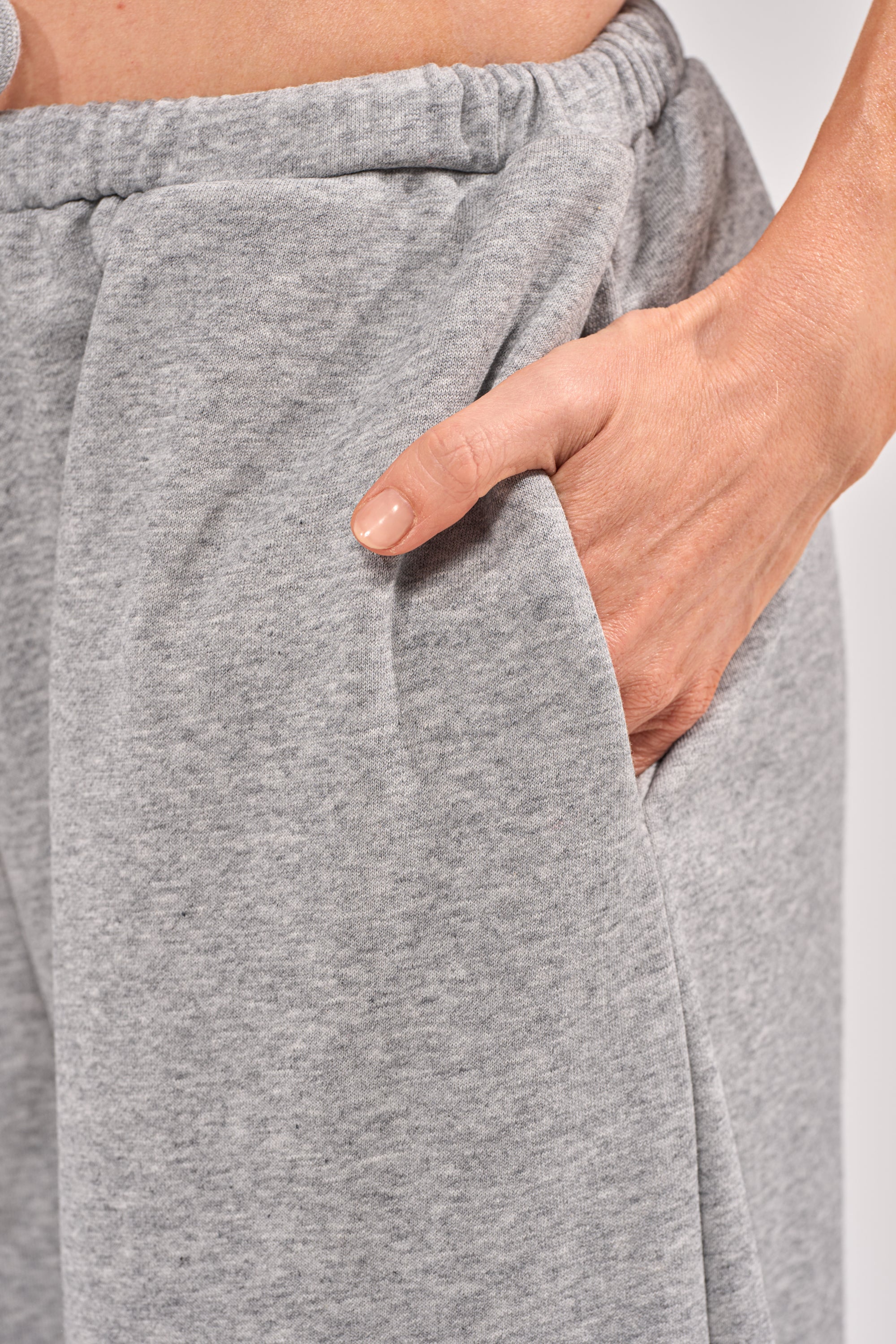 A close-up shows someone wearing CHECK IT OUT GROMMET JOGGER pants in light gray, with their right hand tucked into the pocket. The joggers have a soft, comfy look and an elastic waistband. Their fingernails are short and clean. 6