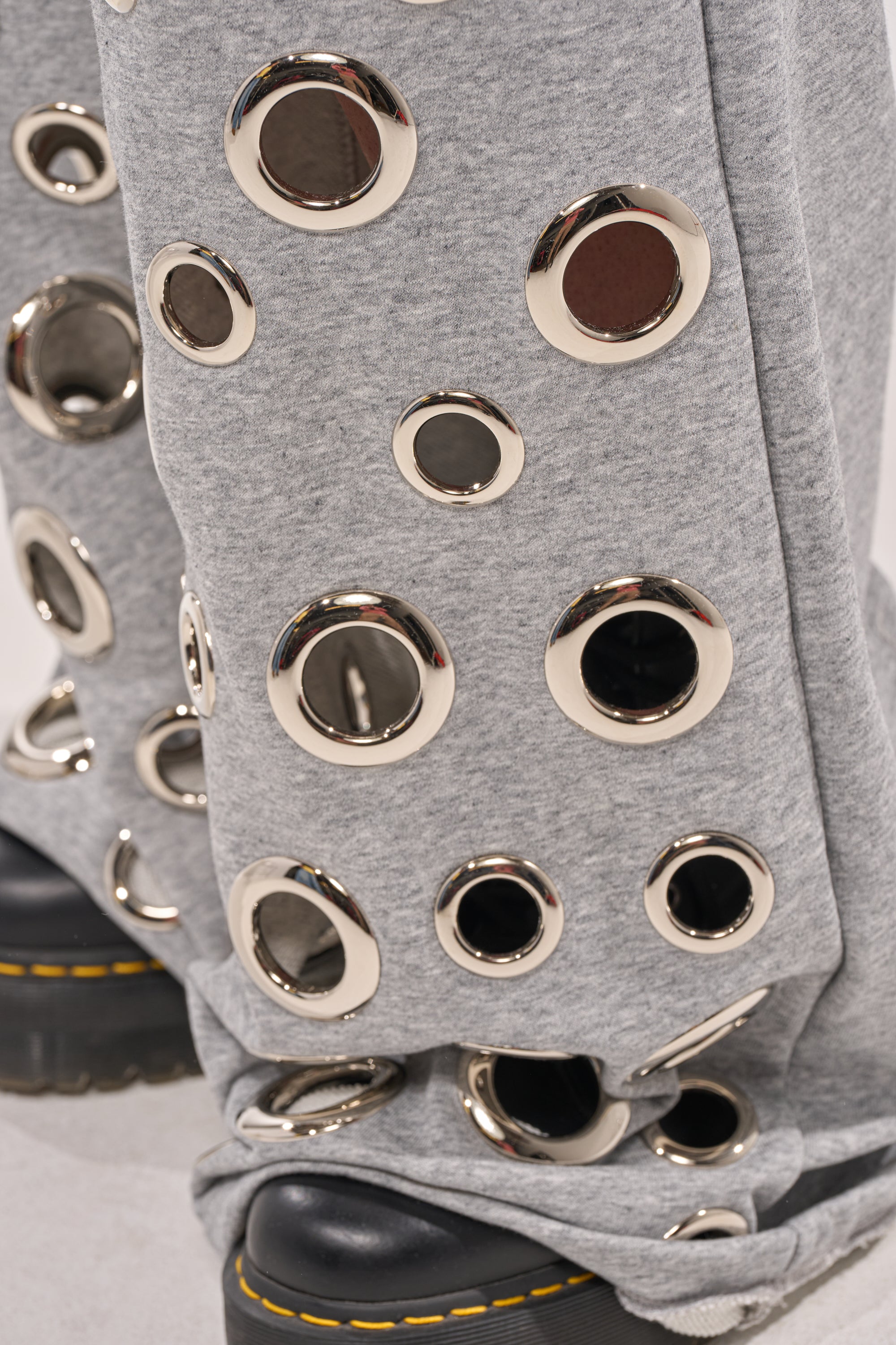 Close-up of the CHECK IT OUT GROMMET JOGGER, grey wide-leg sweats with large silver grommet hardware throughout, styled with black platform shoes featuring yellow stitching. 7