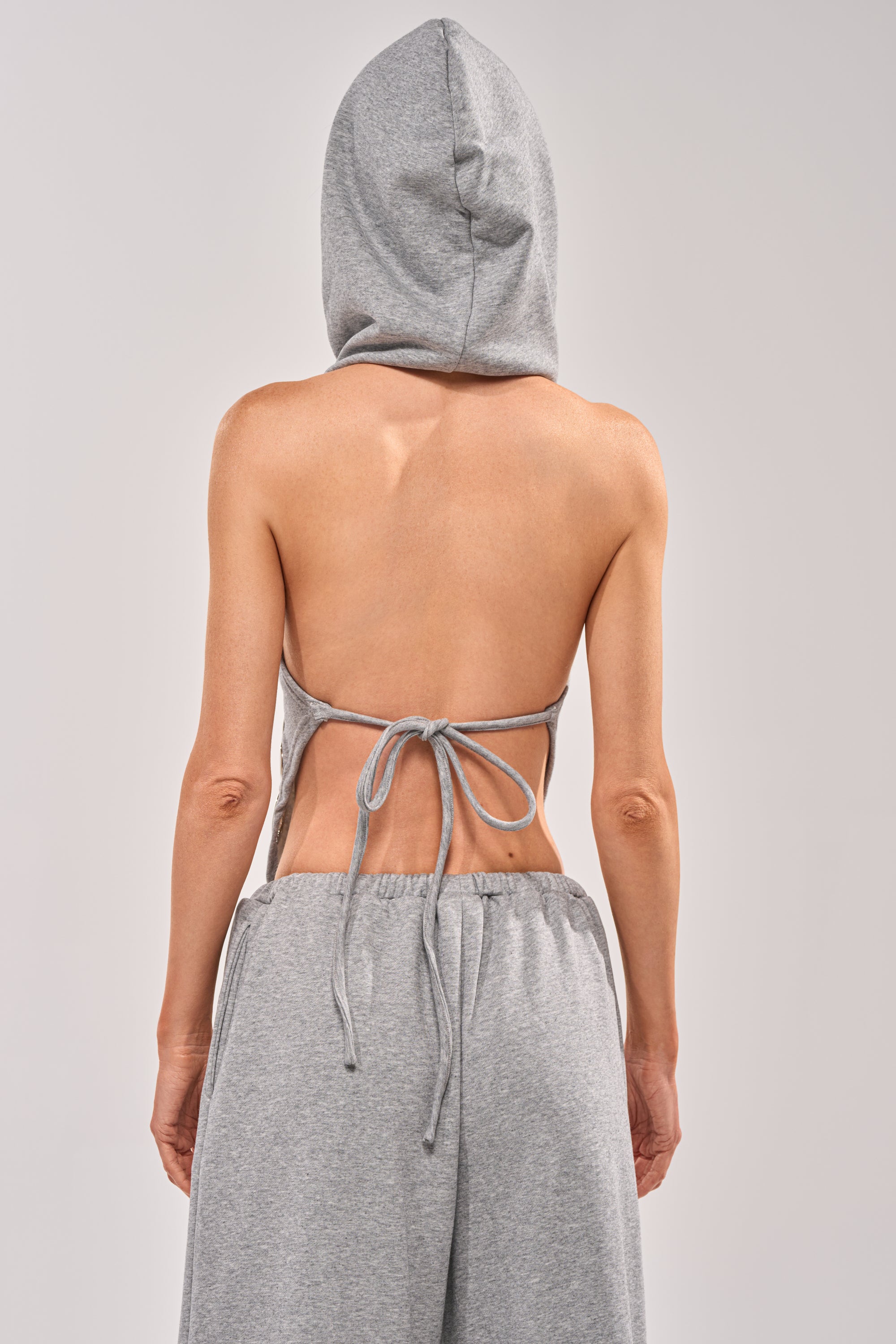 A person stands facing away from the camera, wearing the CHECK IT OUT GROMMET TOP—a gray hooded halter top with grommet detail—and matching grey pants against a plain light background. 5