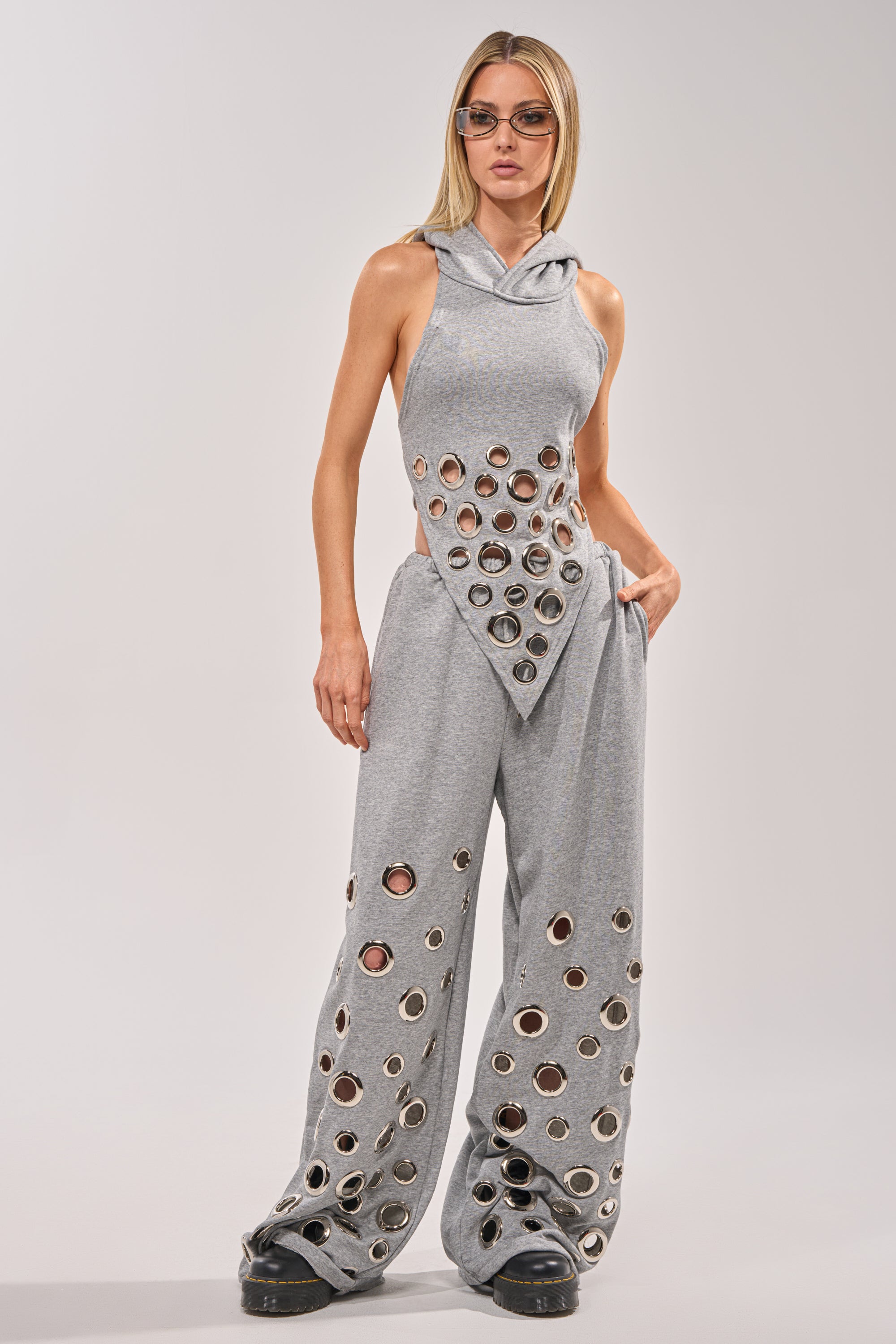 A woman in streetwear rocks the CHECK IT OUT GROMMET TOP, a gray hooded halter with bold grommet details and circular cut-outs. She pairs it with wide-leg pants, sunglasses, black shoes, and has straight blonde hair. 6