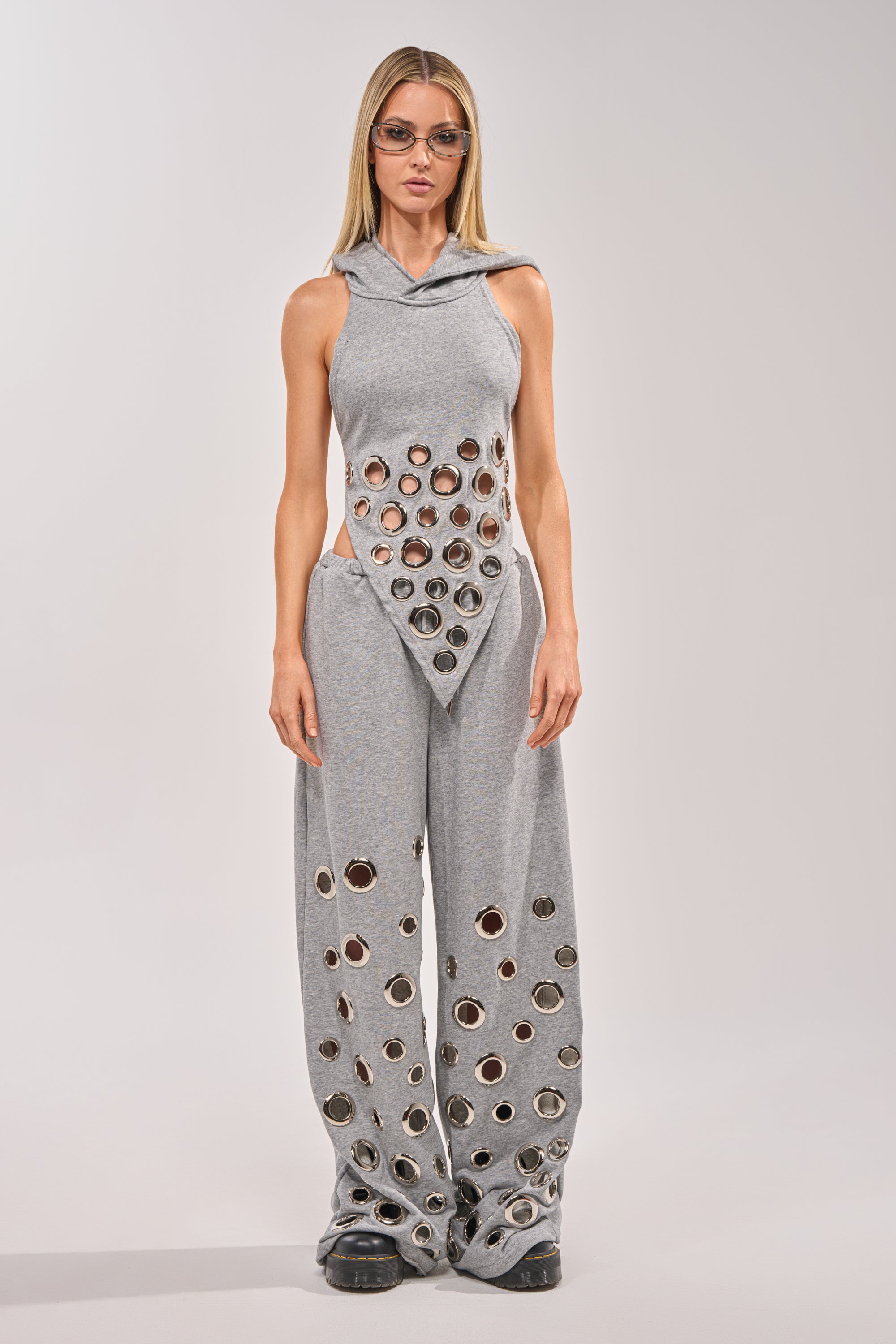 A woman wears the CHECK IT OUT GROMMET TOP, a gray hooded halter with bold circular cutouts and grommet details over red fabric, paired with matching wide-leg pants. She completes her street look with sunglasses and black shoes against a plain background. 7