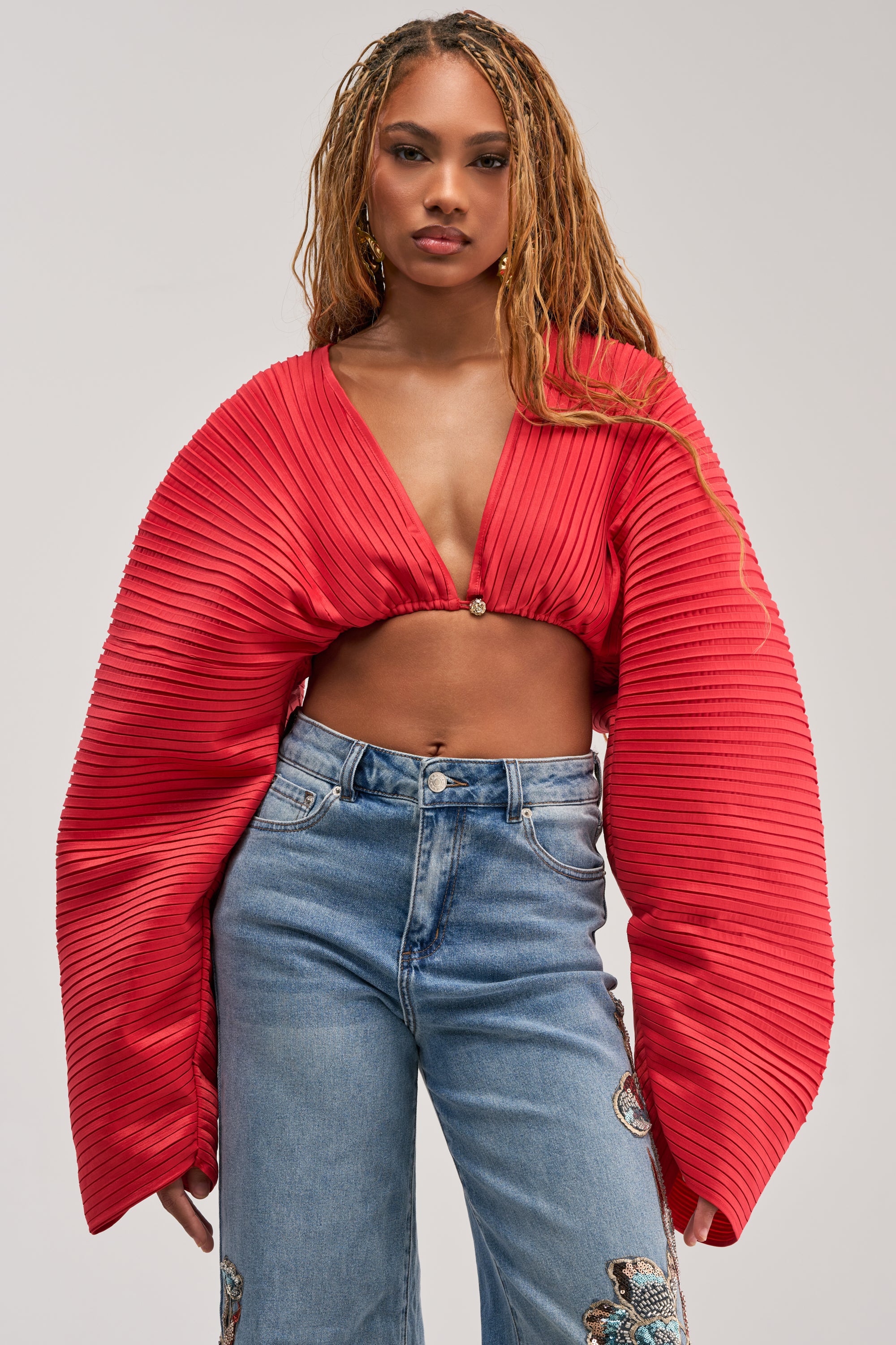 Wearing the FIANA LONG SLEEVE BLOUSE featuring pleated red fabric and wide, balloon sleeves, the woman stands against a plain background paired with high-waisted blue jeans decorated with embroidered floral patches.