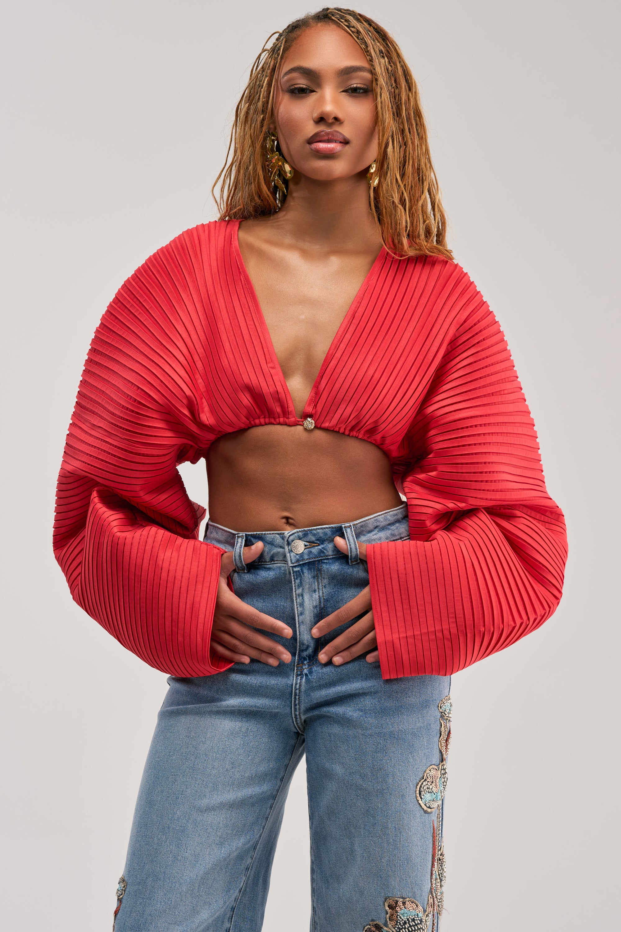 A woman with long braids wears the FIANA LONG SLEEVE BLOUSE—a pleated red top featuring oversized, balloon sleeves—paired with high-waisted blue jeans adorned with embroidery. She stands confidently against a plain background.