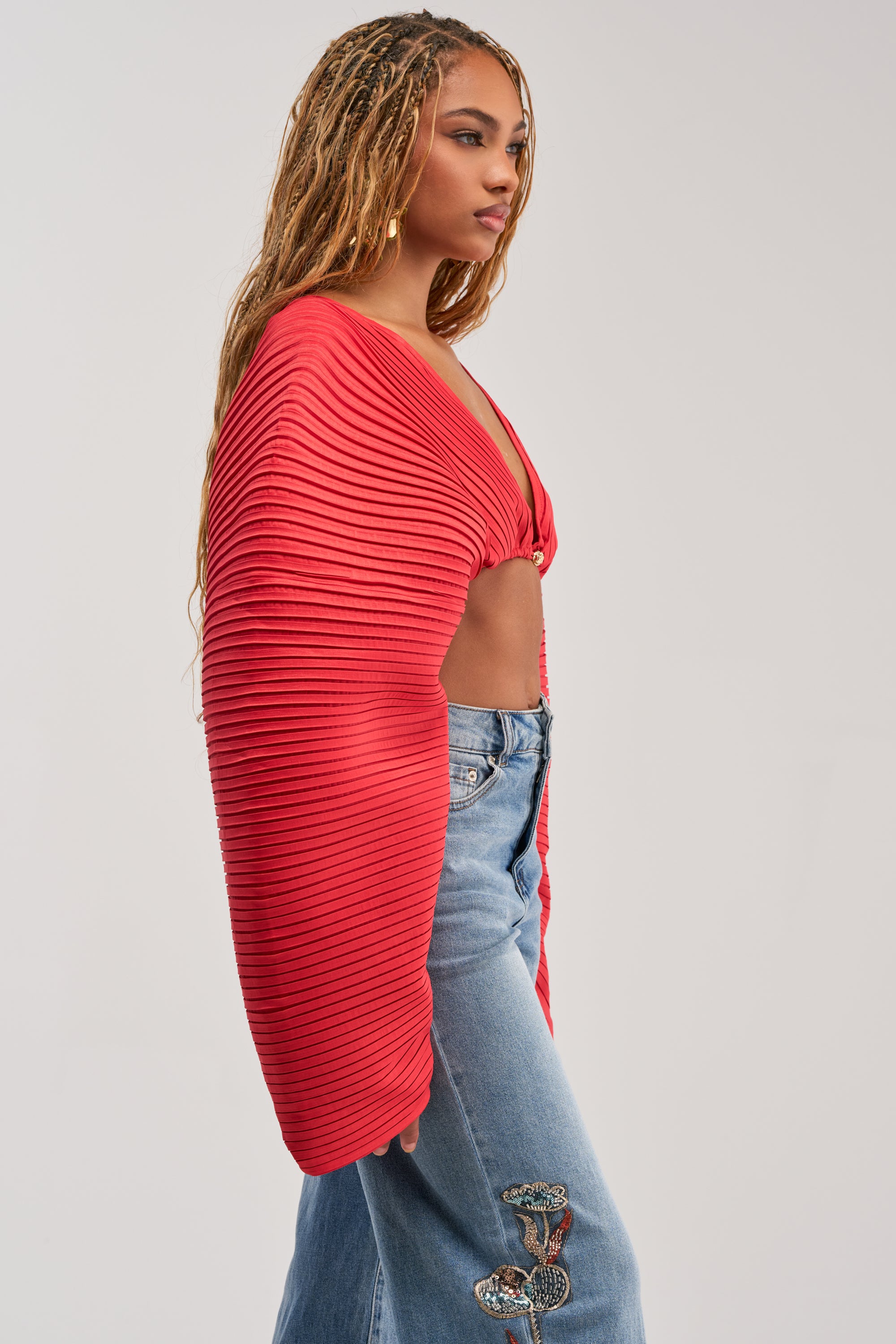 A woman with long braids is seen in profile against a plain background, wearing the FIANA LONG SLEEVE BLOUSE—a pleated red cropped blouse with long sleeves—paired with blue jeans decorated with butterfly patches.