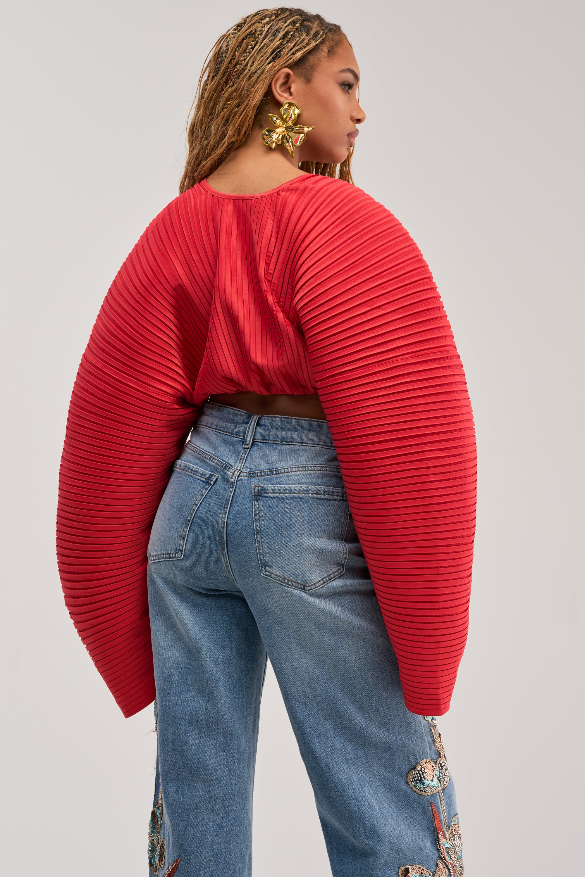 A person wearing the FIANA LONG SLEEVE BLOUSE, featuring pleats and voluminous balloon sleeves in red, pairs it with blue embroidered jeans while standing slightly turned away from the camera to showcase large gold earrings and braided hair.