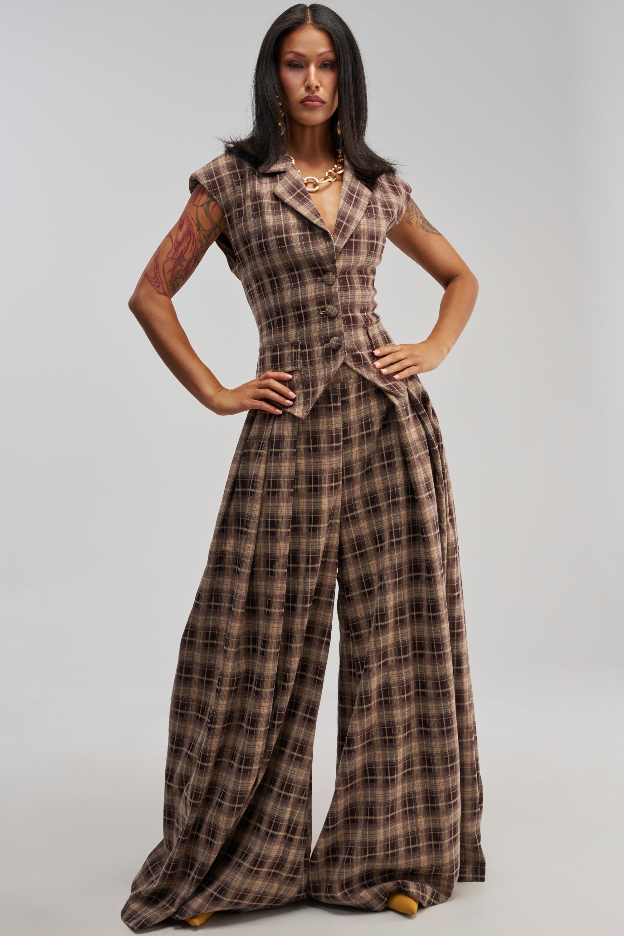 Model showcases the effortlessly chic fall streetwear and workwear styling of the PLAID PRINCESS TROUSER—a sleeveless brown plaid wide-leg jumpsuit—paired with a gold necklace and yellow shoes, posing confidently with hands on hips against a plain background.